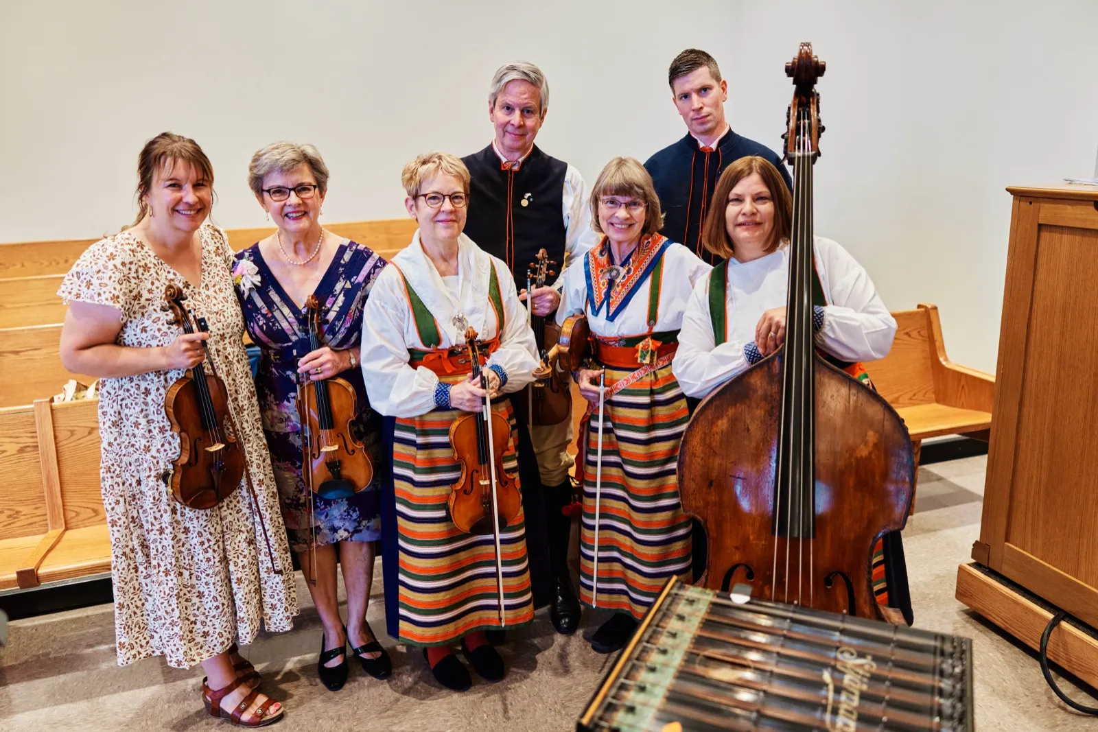 A group of seven folk musicians in traditional Scandinavian attire pose with violins, an upright bass, and a hammered dulcimer inside a chapel. — Tim Larsen Photography, Minnesota