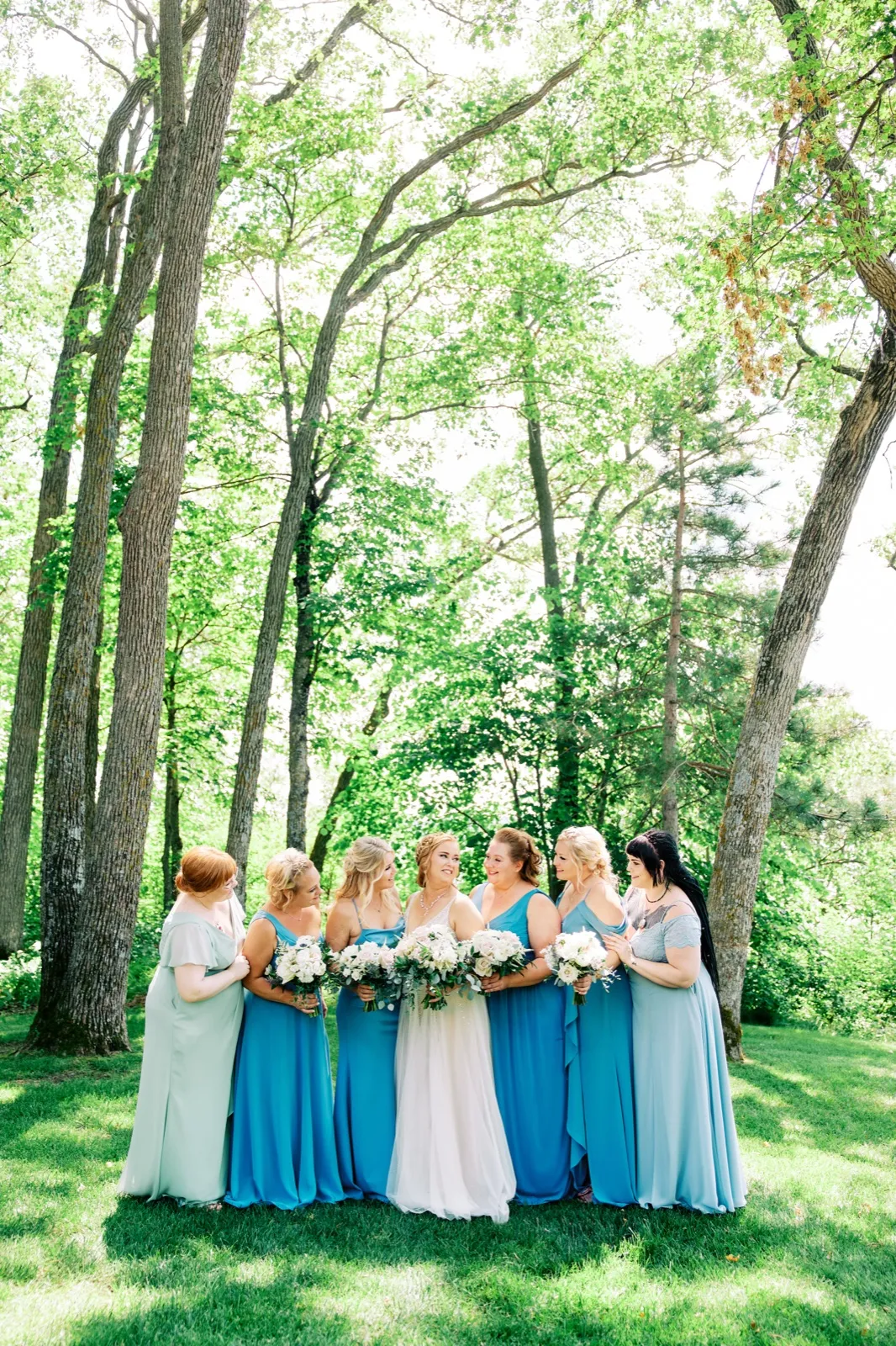 The bride stands centered with seven bridesmaids in varying shades of blue dresses, all holding white bouquets, beneath towering trees on a sunlit lawn. — Tim Larsen Photography, Brainerd Lakes MN