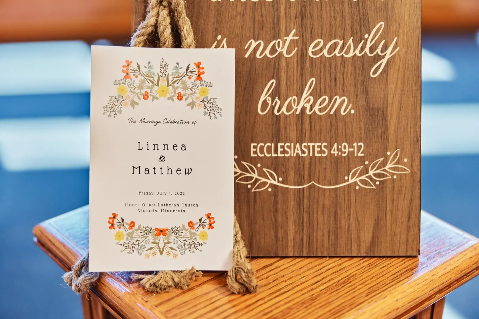 A floral wedding program for Linnea and Matthew leans against a wooden sign engraved with the Ecclesiastes 4:9-12 verse on a wooden bench. — Tim Larsen Photography, Minnesota