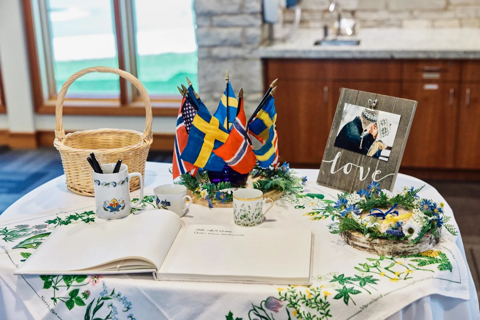 A guest book table displays an open signing book, Scandinavian flags from Norway and Sweden, heritage china mugs, a love photo frame, and floral arrangements on a botanical tablecloth. — Tim Larsen Photography, Minnesota