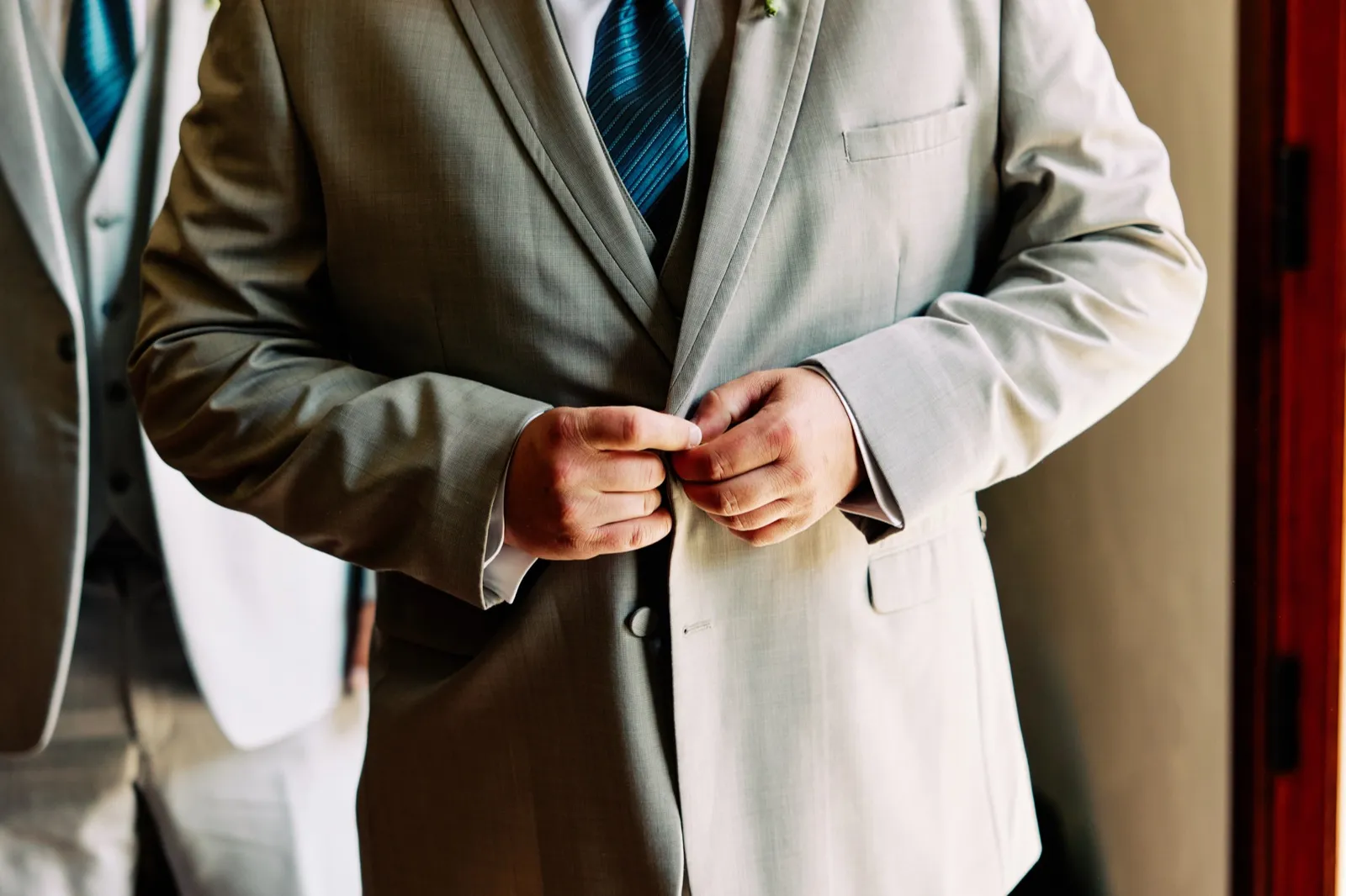 Close-up of the groom's hands buttoning his tan suit jacket over a blue striped tie while a groomsman stands beside him. — Tim Larsen Photography, Brainerd Lakes MN