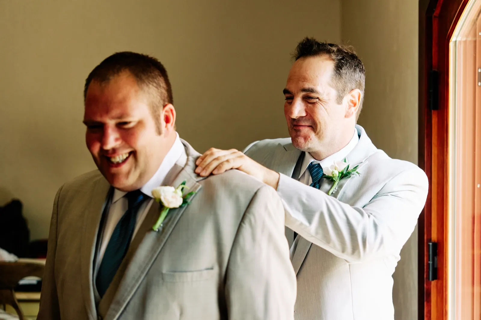 A groomsman smooths the groom's tan suit jacket from behind as the groom laughs in warm window light. — Tim Larsen Photography, Brainerd Lakes MN