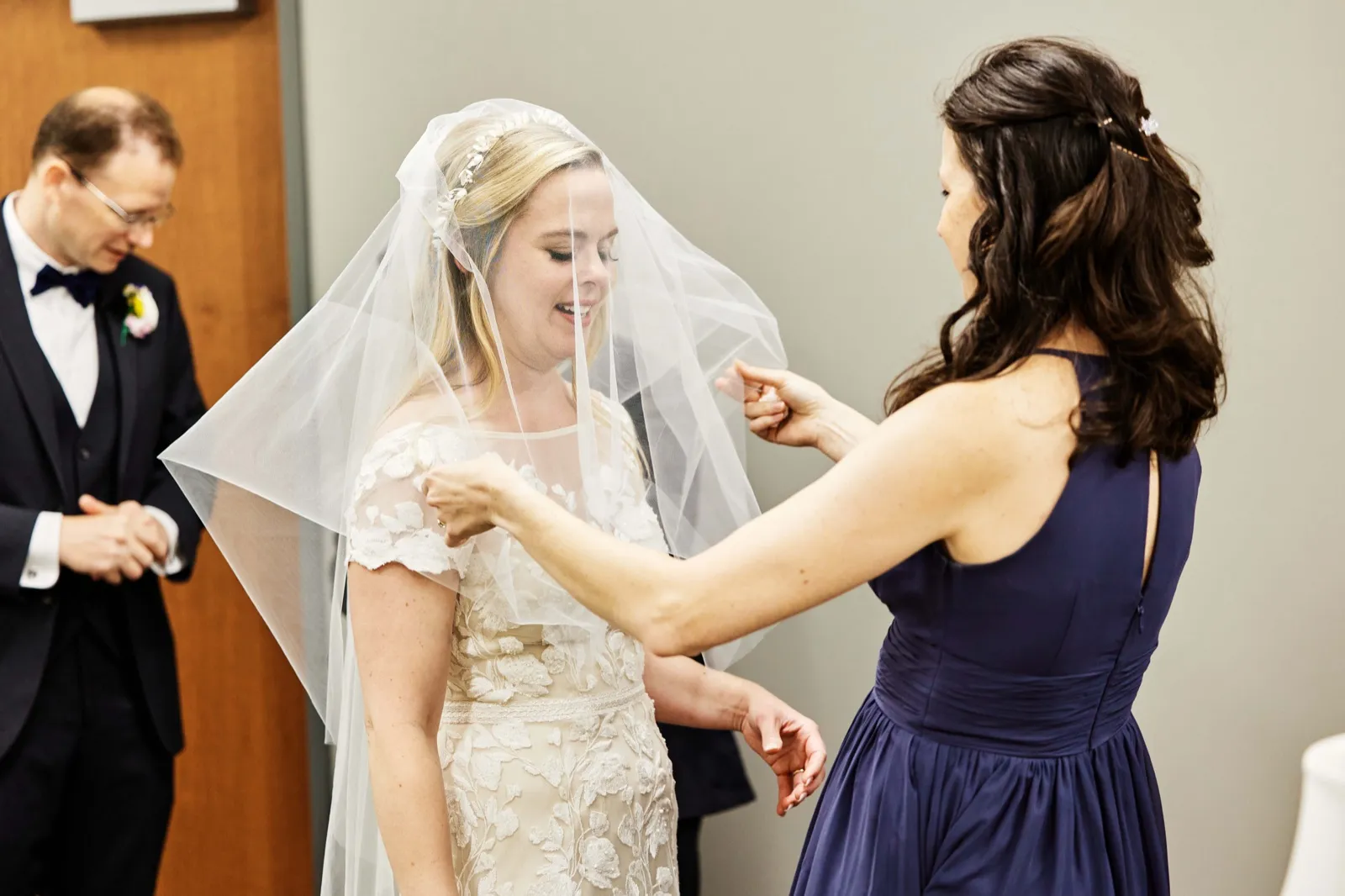 An attendant in a navy dress arranges the bride's veil over her face as the bride laughs, with the groom visible out of focus in the background. — Tim Larsen Photography, Minnesota