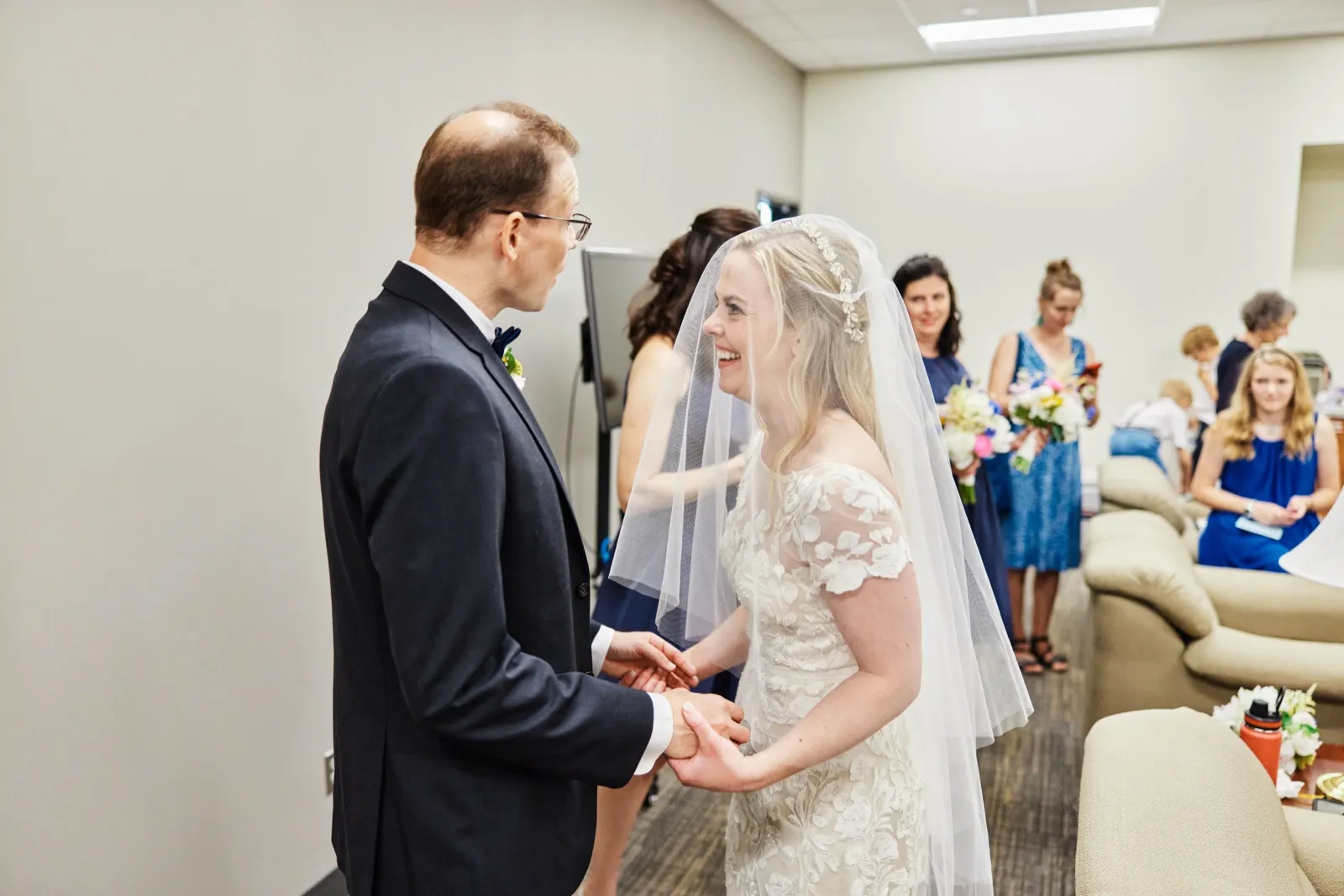 The bride laughs joyfully while holding the groom's hands during a first look in a casual indoor room with the bridal party gathered behind. — Tim Larsen Photography, Minnesota