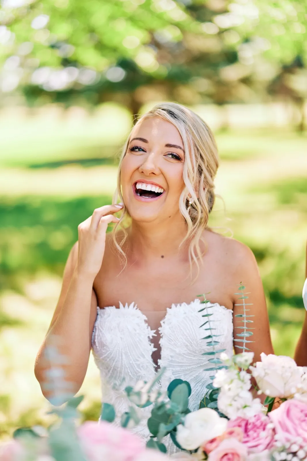 The bride laughs openly with her hand near her chin, holding a pastel bouquet of roses and eucalyptus on a sunlit lawn. — Tim Larsen Photography, Twin Cities MN