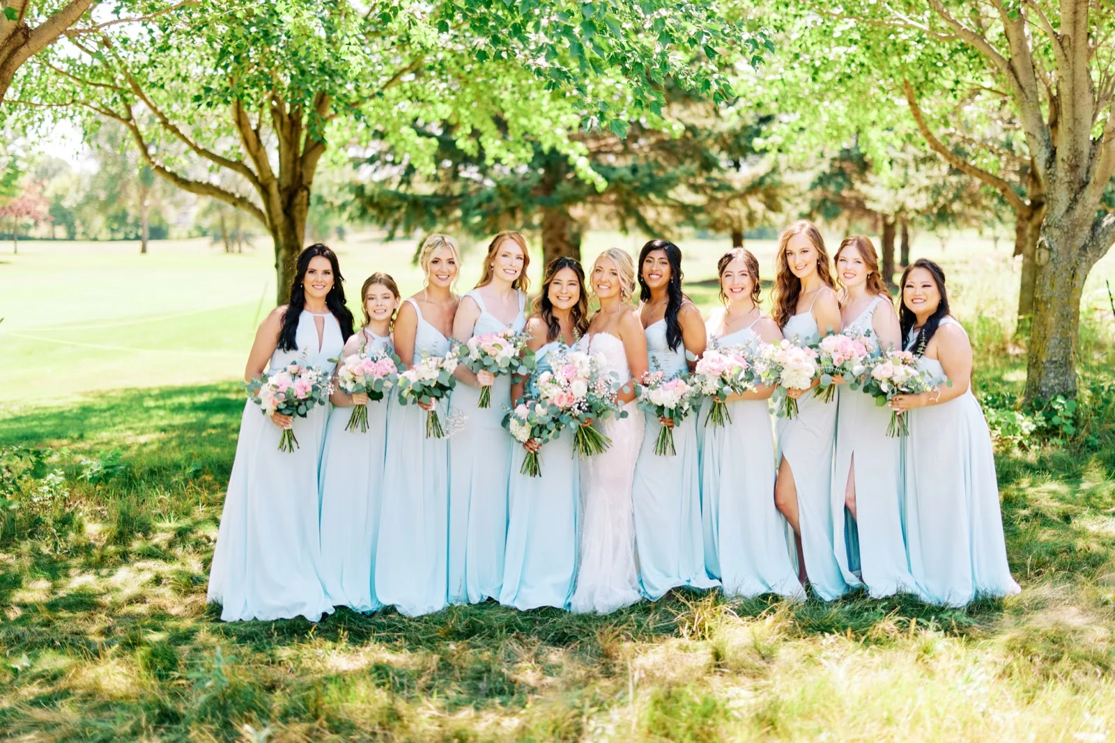 The bride stands at the center of eleven bridesmaids in pale blue gowns, all holding pink and white bouquets, on a sunlit lawn beneath leafy trees. — Tim Larsen Photography, Twin Cities MN