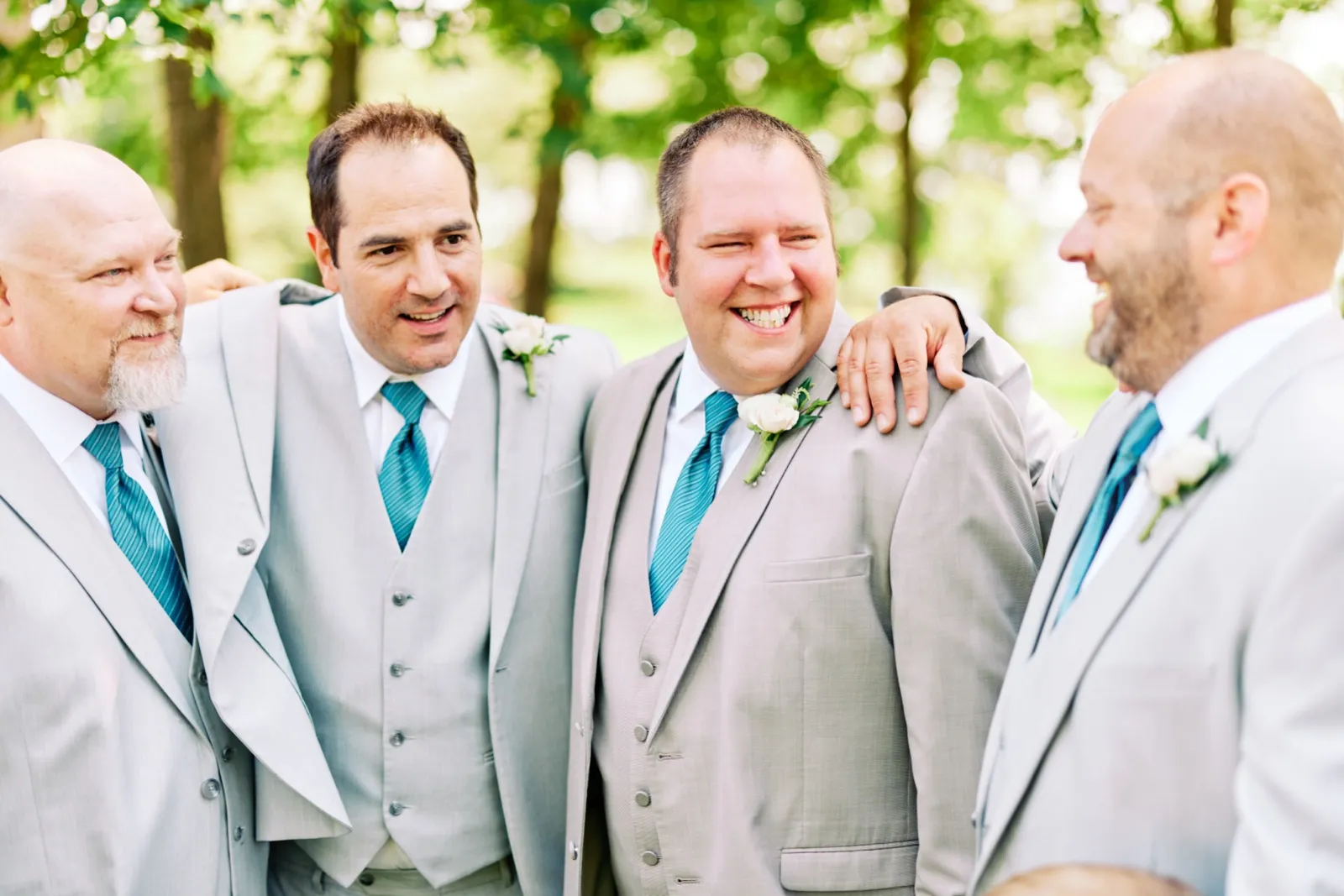 The groom and three groomsmen in gray suits with teal ties laugh together, arms around each other's shoulders, in front of a soft green tree backdrop. — Tim Larsen Photography, Brainerd Lakes MN