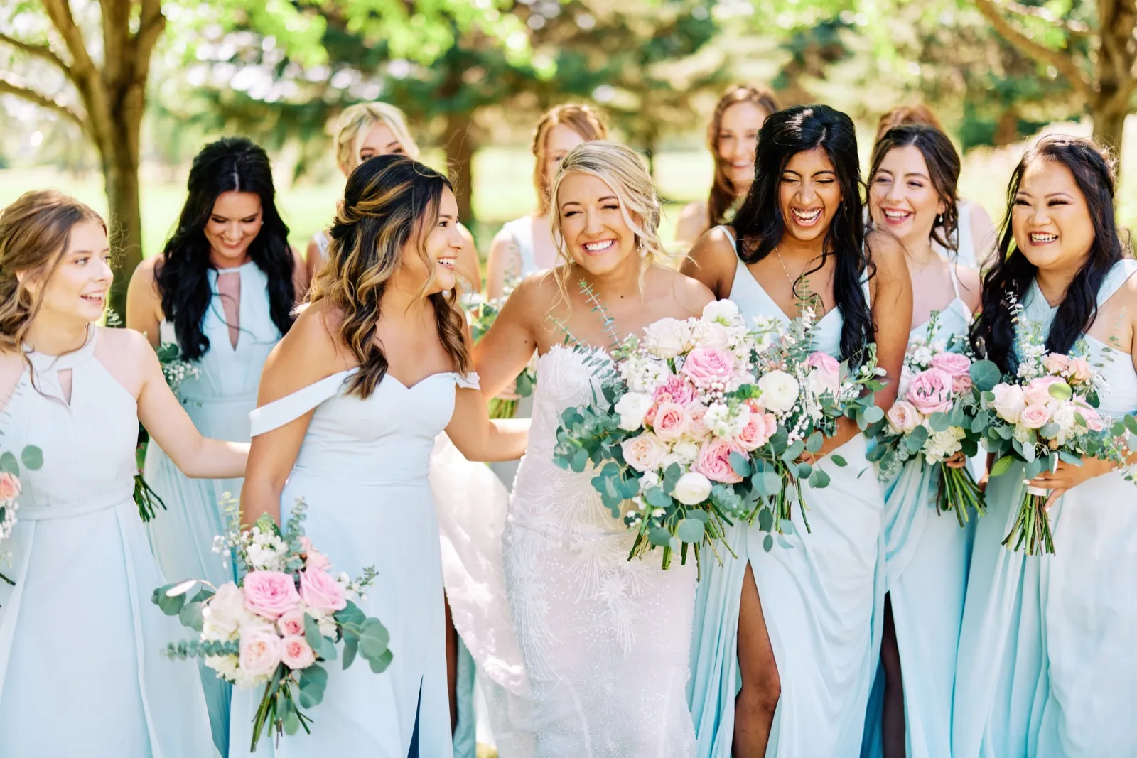 The bride laughs in the center of her bridesmaids, who are dressed in dusty blue gowns and holding pink and white bouquets, on a sunlit lawn. — Tim Larsen Photography, Twin Cities MN