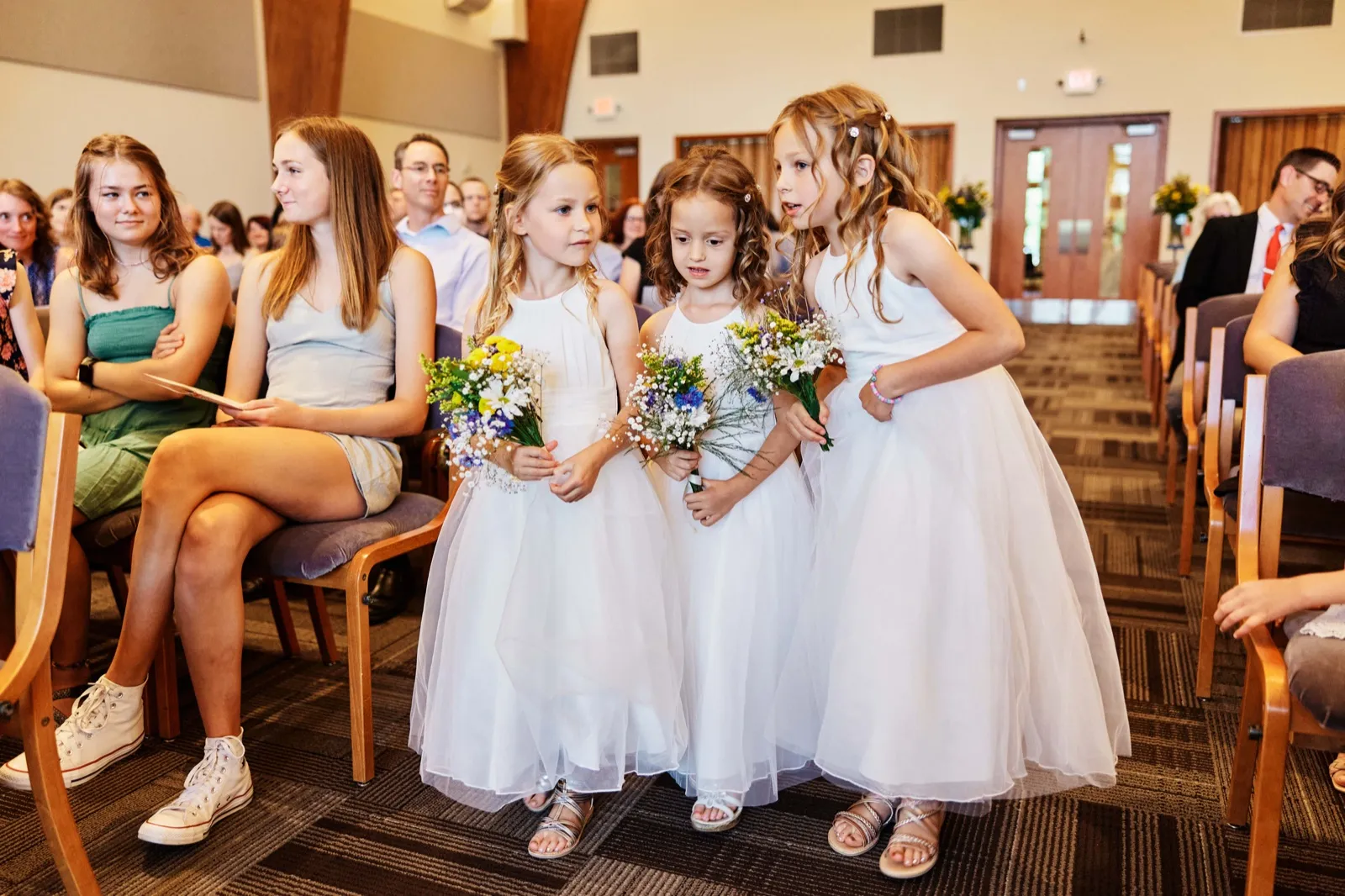 Three flower girls in white dresses pause mid-aisle holding wildflower bouquets, with seated guests visible on the left. — Tim Larsen Photography, Minnesota