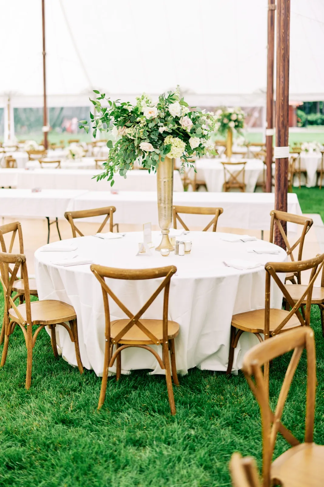 A round reception table with white linen and a tall gold vase holding a lush greenery and blush floral centerpiece, surrounded by wooden cross-back chairs under a sailcloth tent on a lakeside lawn. — Tim Larsen Photography, Brainerd Lakes MN