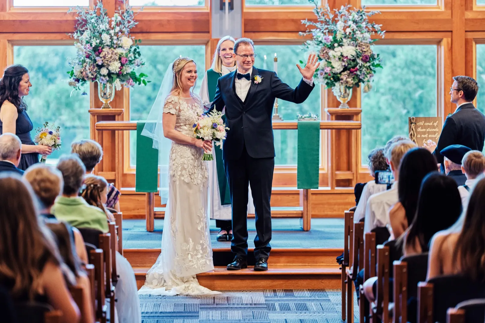 The groom waves toward guests as he and the smiling bride stand at the altar with the officiant behind them, framed by tall floral arrangements and large windows. — Tim Larsen Photography, Minnesota