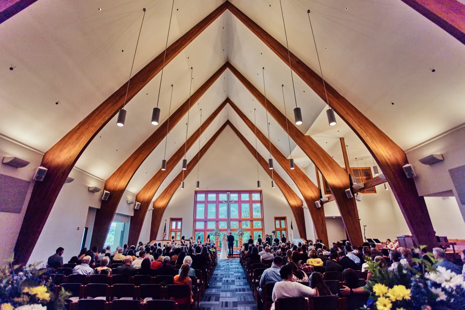 An extreme wide view of a wedding ceremony shows the couple at a glass-walled altar beneath a soaring wood-arched chapel ceiling, with a full congregation seated in rows on either side of the aisle. — Tim Larsen Photography, Minnesota