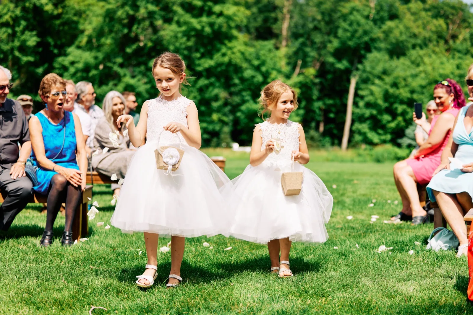 Two flower girls in white dresses walk down a grass aisle holding petal baskets while seated guests watch on either side. — Tim Larsen Photography, Brainerd Lakes MN