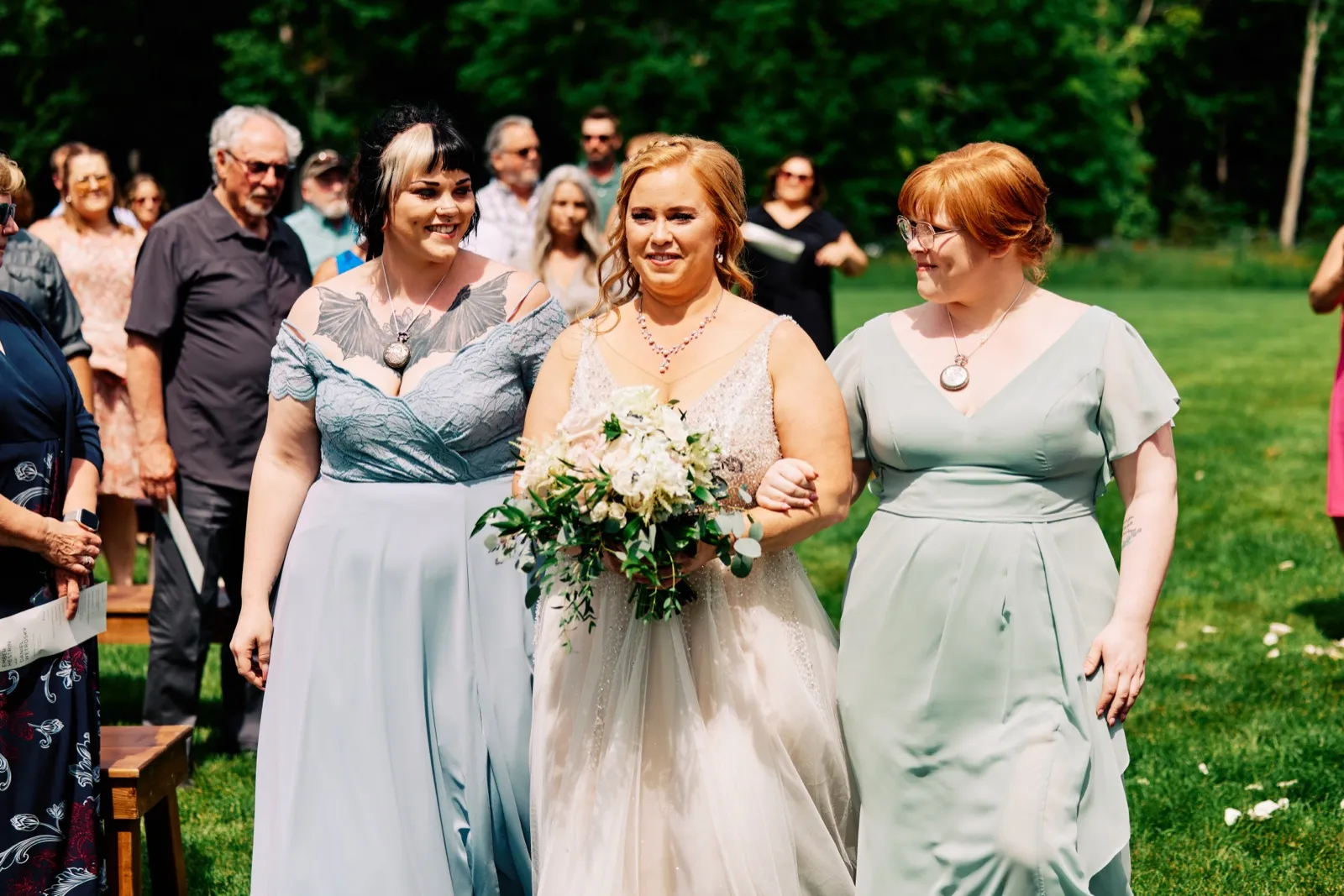 The bride walks down a grass aisle flanked by two attendants in dusty blue and sage dresses, holding a white and green bouquet as guests look on. — Tim Larsen Photography, Brainerd Lakes MN