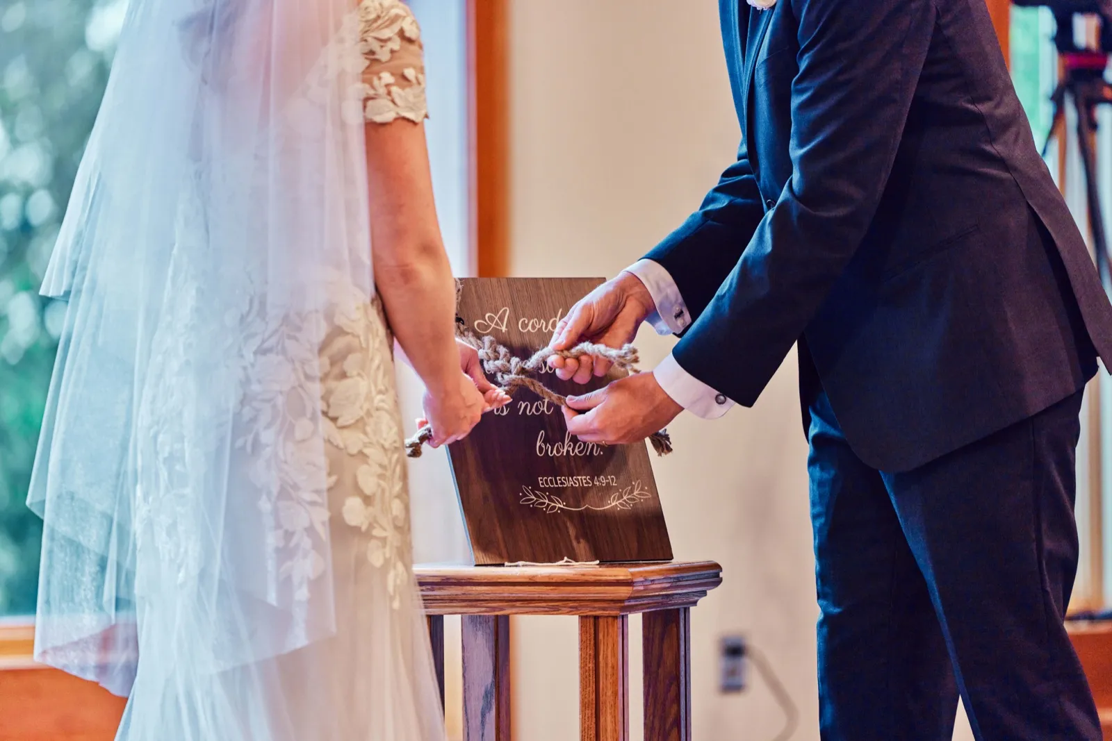 The bride and groom braid a three-strand cord together during their ceremony, with the Ecclesiastes 4:9-12 sign visible behind their hands. — Tim Larsen Photography, Minnesota