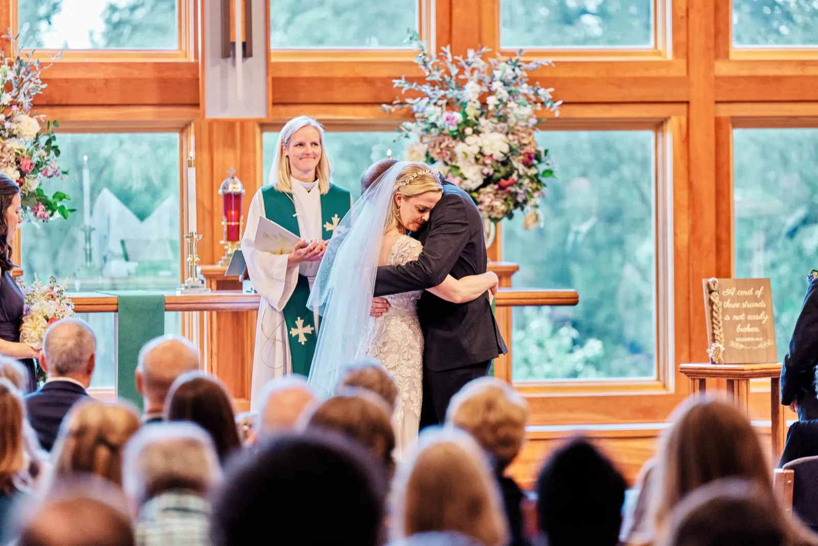 The bride and groom embrace at the altar during their ceremony as a smiling officiant looks on, framed by tall windows and floral arrangements. — Tim Larsen Photography, Minnesota