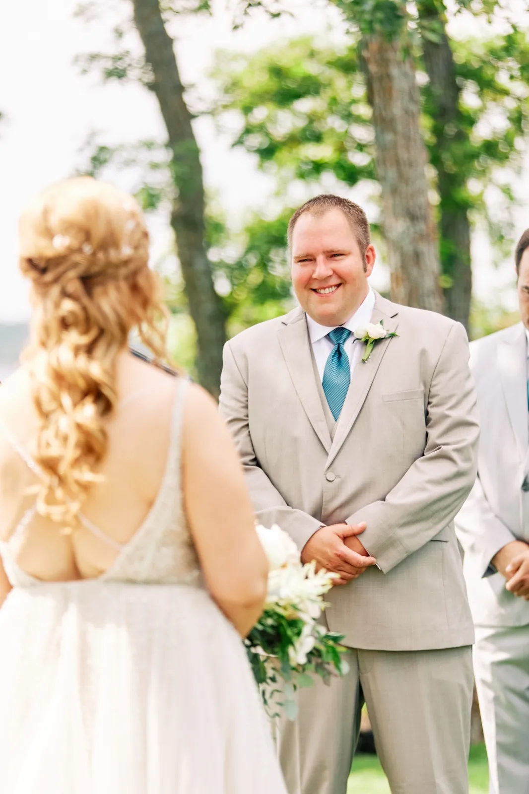 The groom smiles broadly at the bride during their lakeside ceremony, framed over her shoulder with her bouquet visible in the foreground. — Tim Larsen Photography, Brainerd Lakes MN