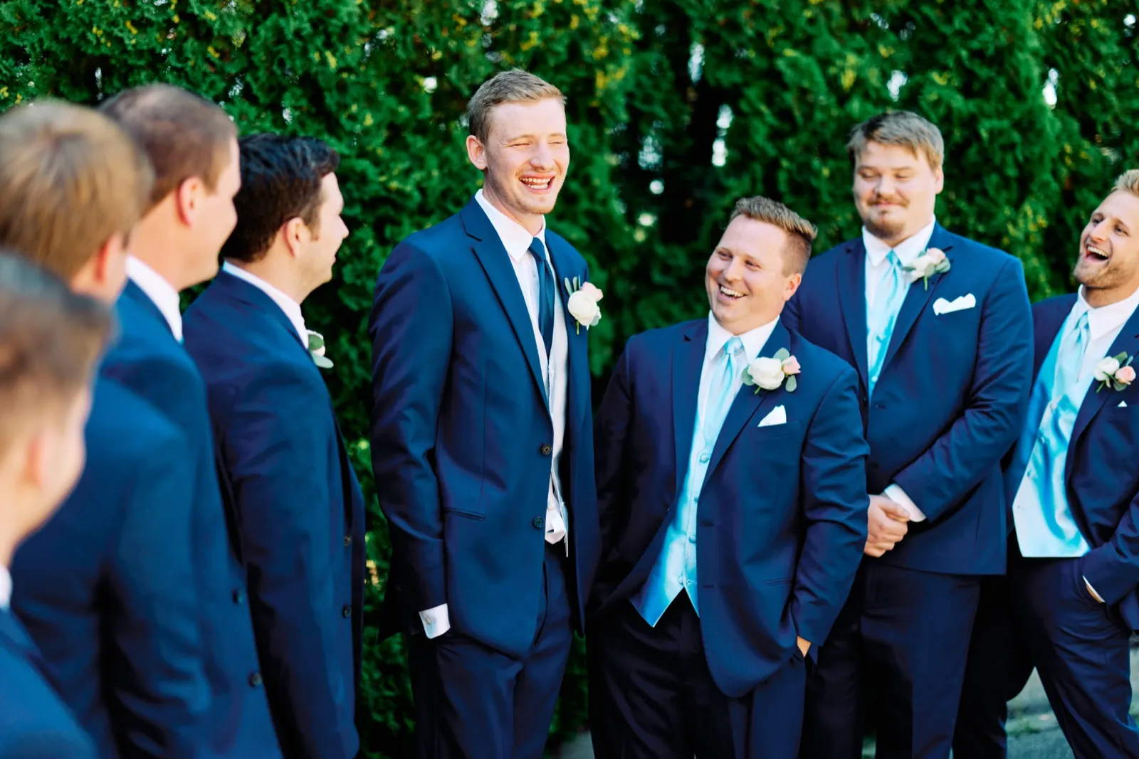 The groom laughs in the center of his groomsmen, all in navy suits with light blue ties, against a tall green hedge. — Tim Larsen Photography, Twin Cities MN