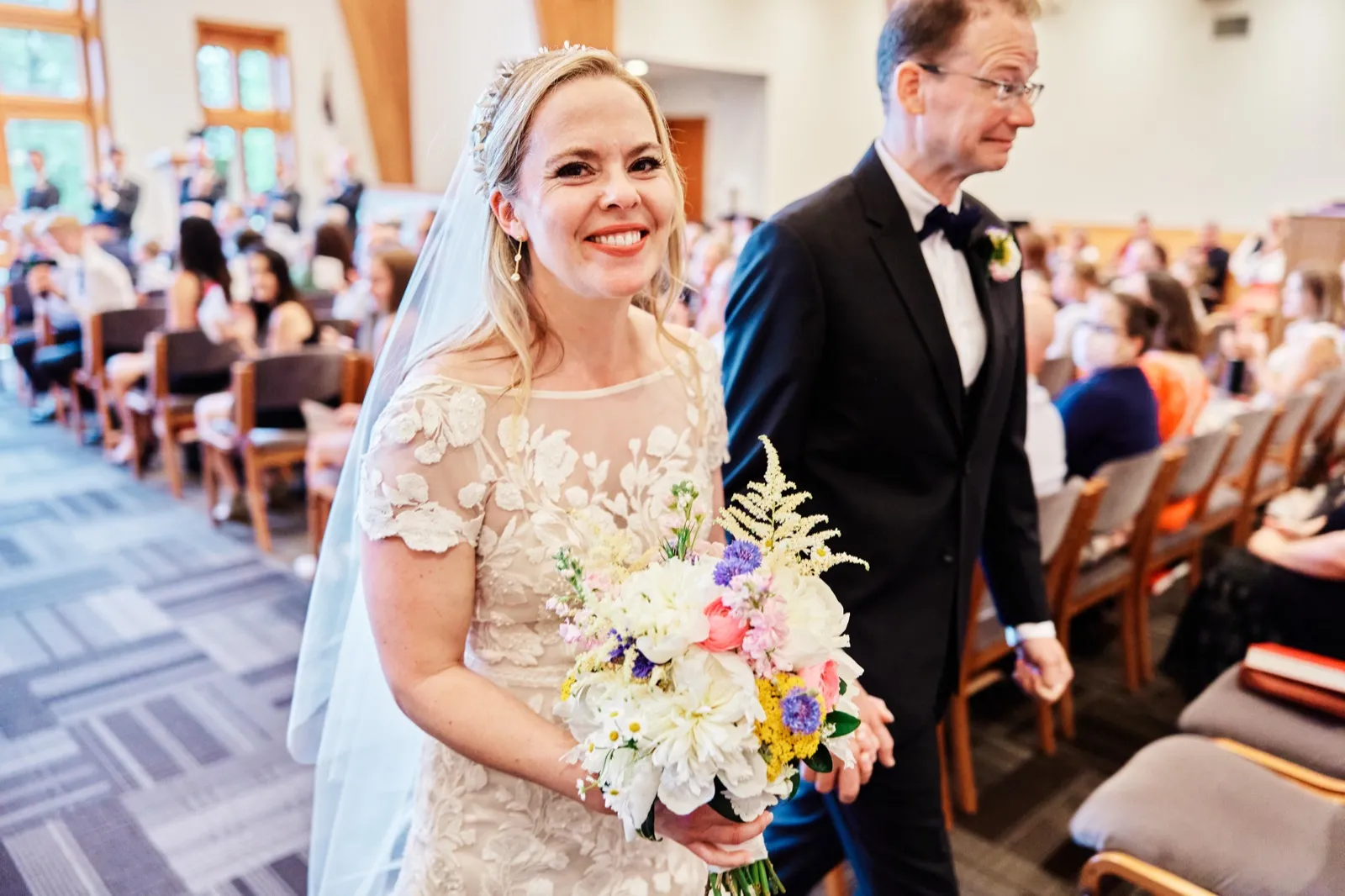 The bride beams as she walks down the ceremony aisle holding a pastel bouquet, her father in a tuxedo at her side and guests filling the pews behind them. — Tim Larsen Photography, Minnesota