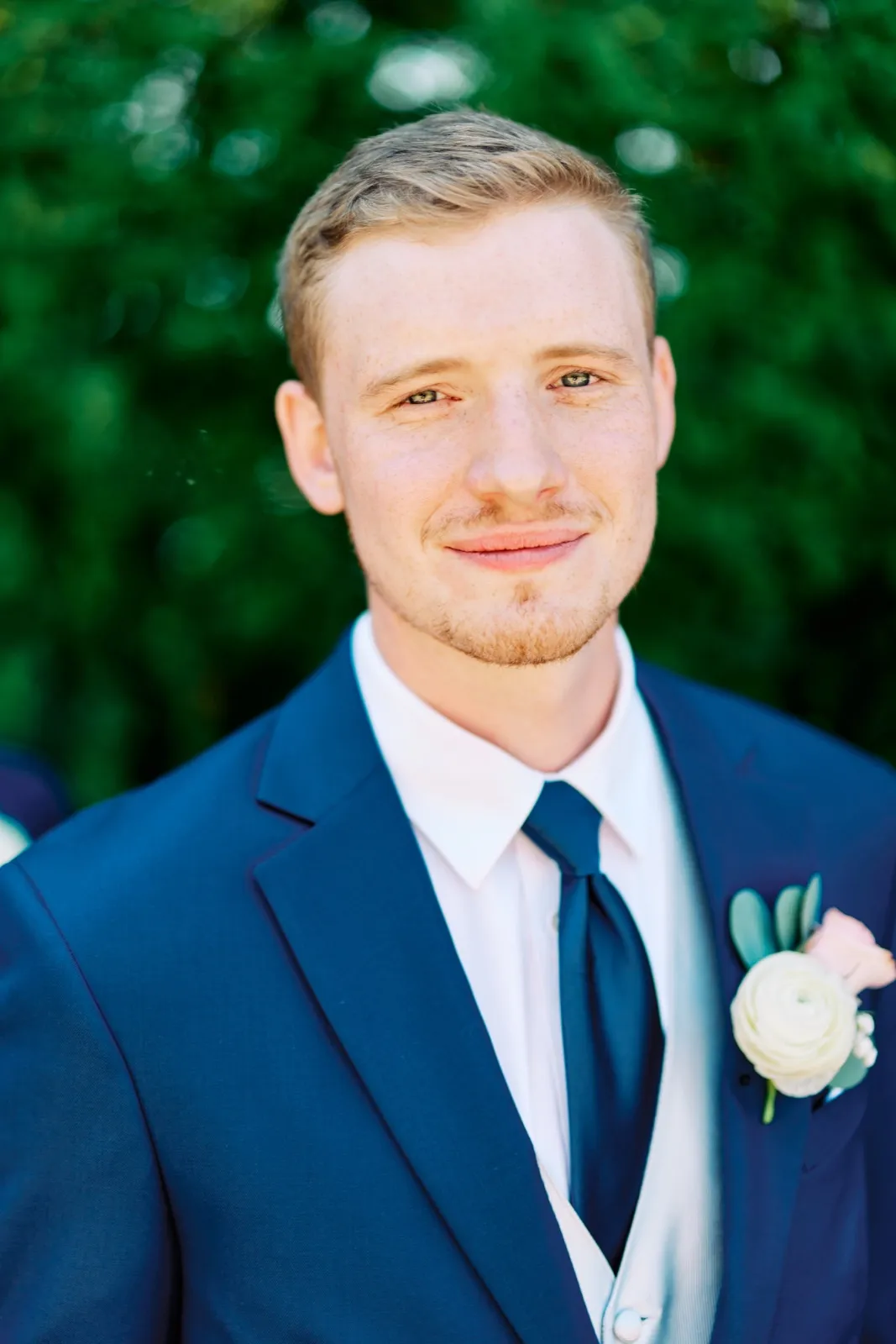 The groom in a navy suit and tie with a white ranunculus boutonniere stands against a green hedge, looking softly toward the camera. — Tim Larsen Photography, Twin Cities MN
