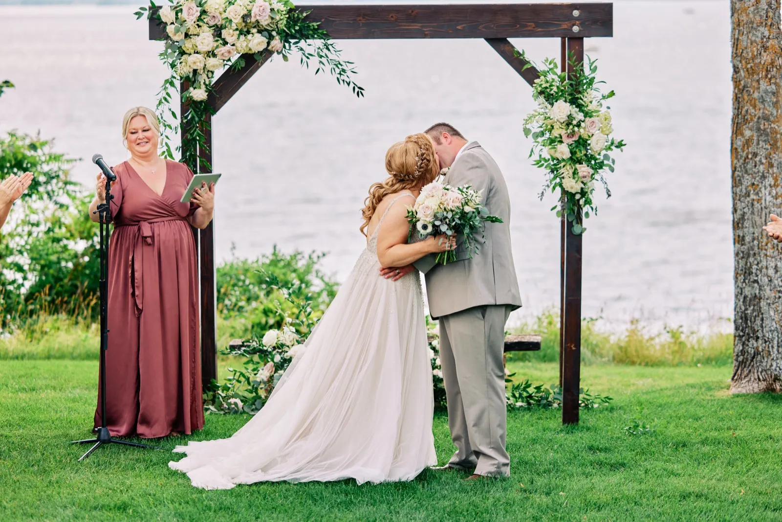 The bride and groom share a kiss beneath a wooden floral arch as the smiling officiant looks on, with Gull Lake stretching behind them. — Tim Larsen Photography, Brainerd Lakes MN