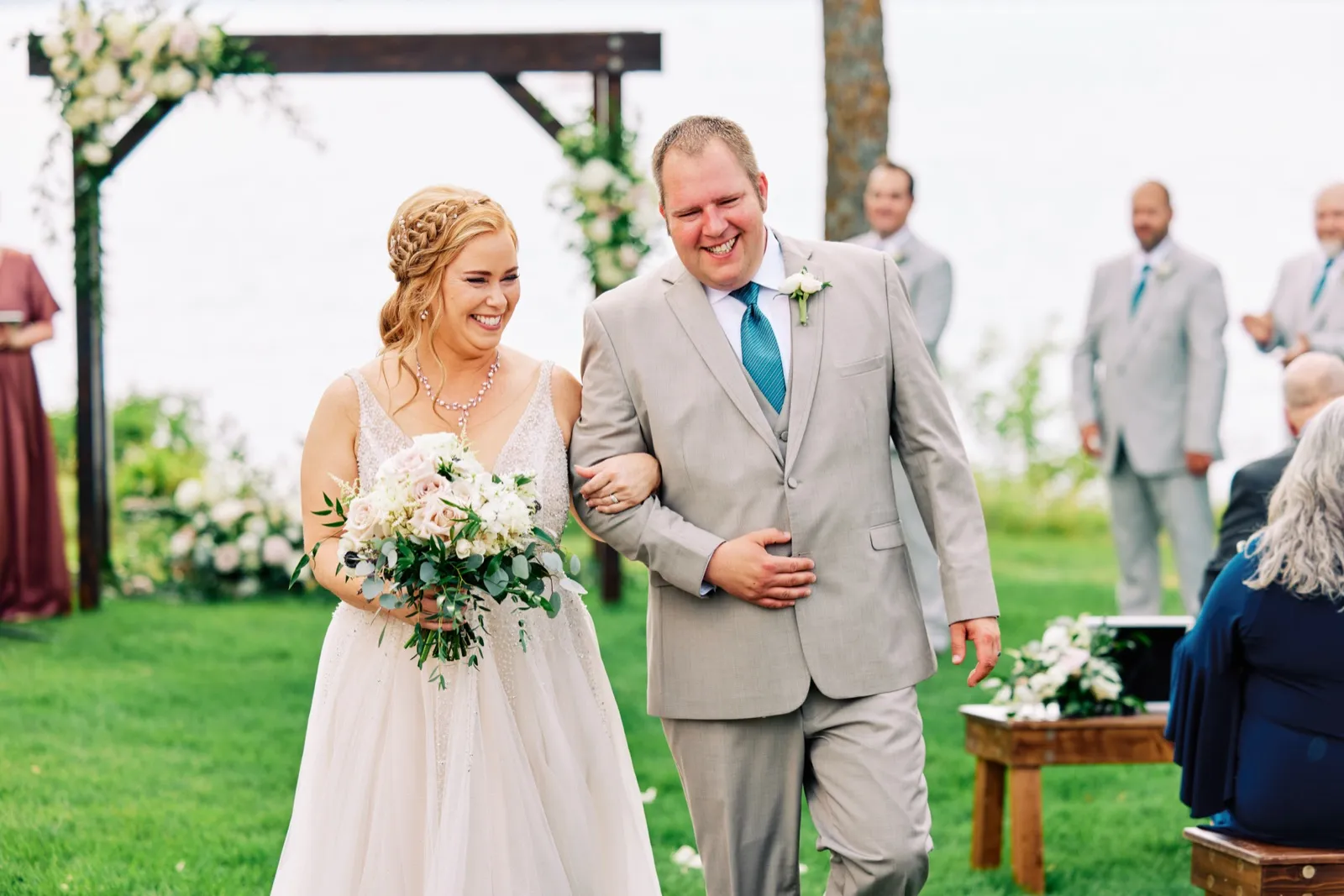 The bride and groom laugh as they walk back down the aisle together after their lakeside ceremony, the bride holding a soft pastel bouquet. — Tim Larsen Photography, Brainerd Lakes MN
