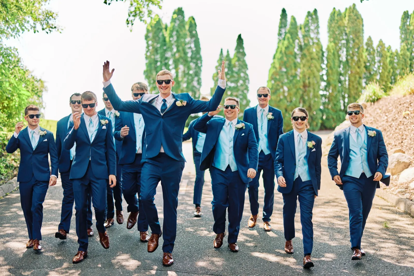 The groom walks down a tree-lined path with arms raised triumphantly, flanked by ten groomsmen in navy suits and sunglasses. — Tim Larsen Photography, Twin Cities MN