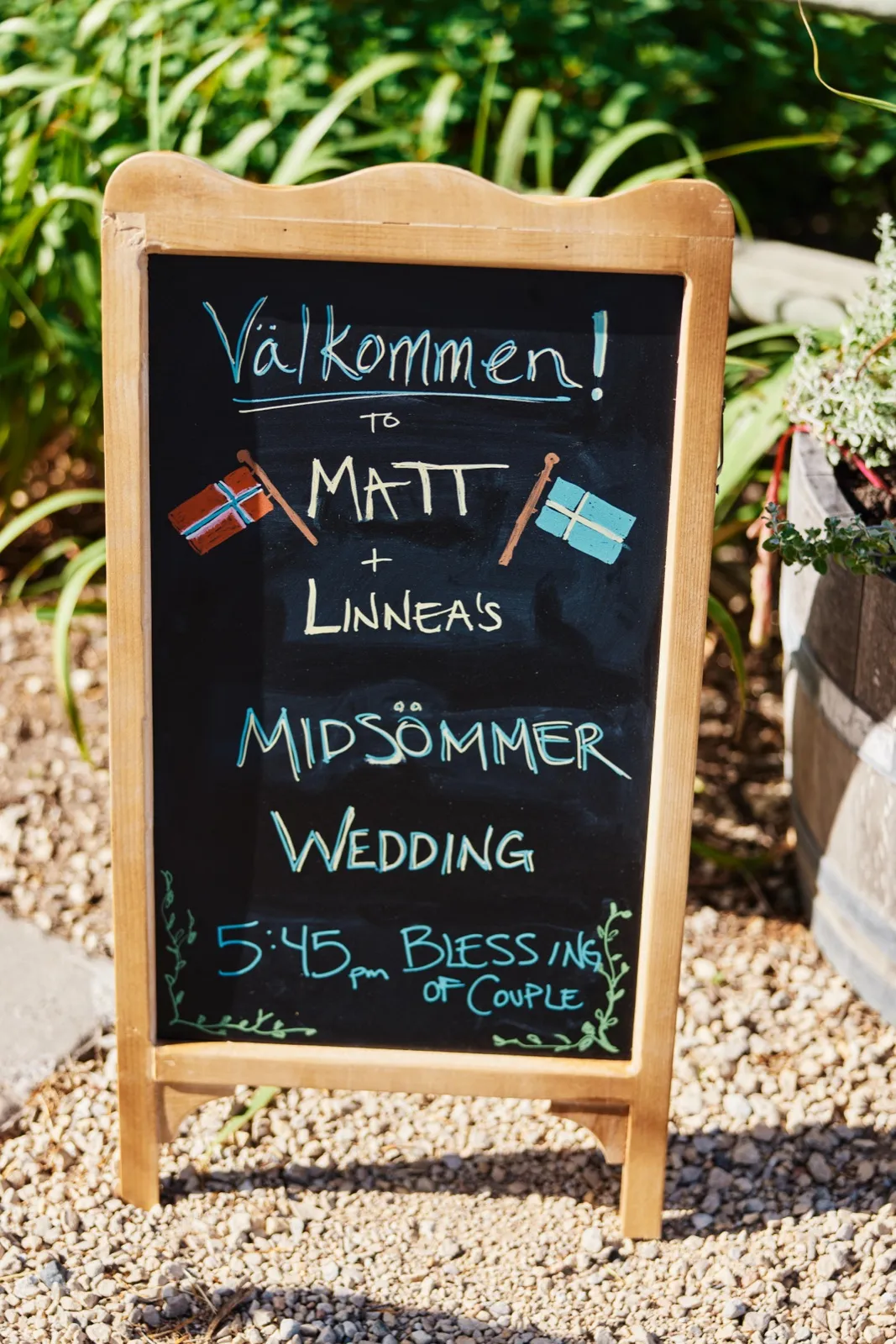 A handwritten chalkboard sign welcomes guests to Matt and Linnea's Midsommer Wedding with Norwegian and Swedish flags drawn above the names and a 5:45 pm blessing of the couple noted at the bottom. — Tim Larsen Photography, Minnesota