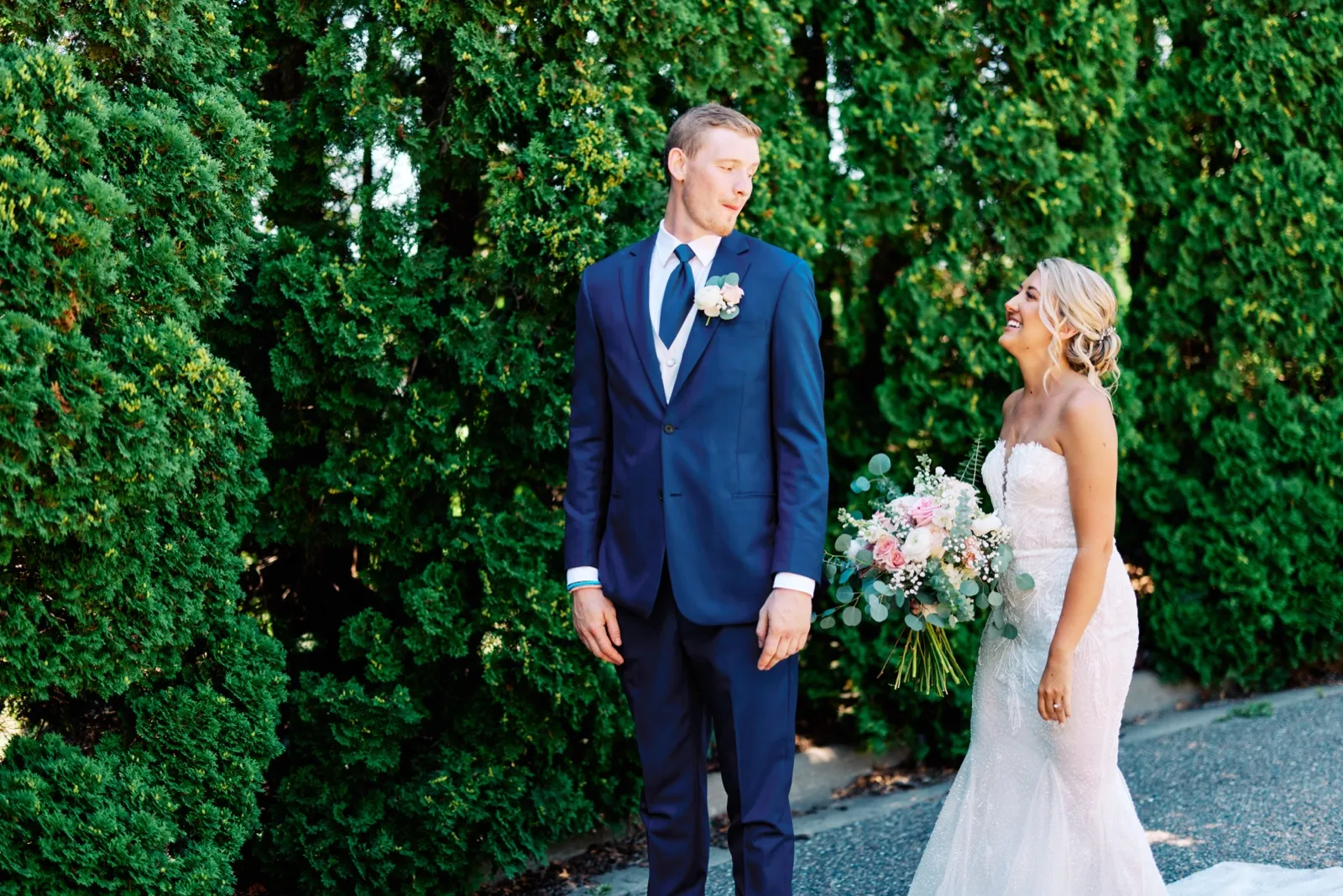 The bride laughs while looking at the groom on a garden path framed by a tall green arborvitae hedge, holding a pink and white bouquet. — Tim Larsen Photography, Twin Cities MN
