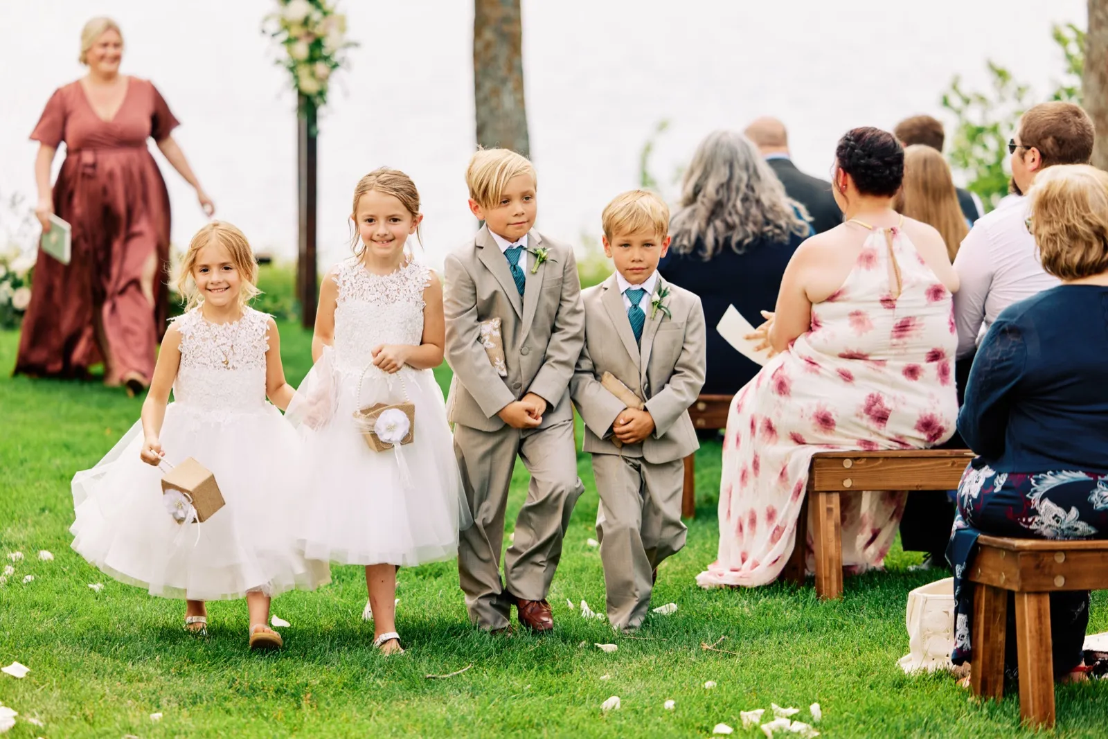 Two flower girls in white dresses and two ring bearers in gray suits walk down a grass aisle while seated guests look on at a lakeside ceremony. — Tim Larsen Photography, Brainerd Lakes MN