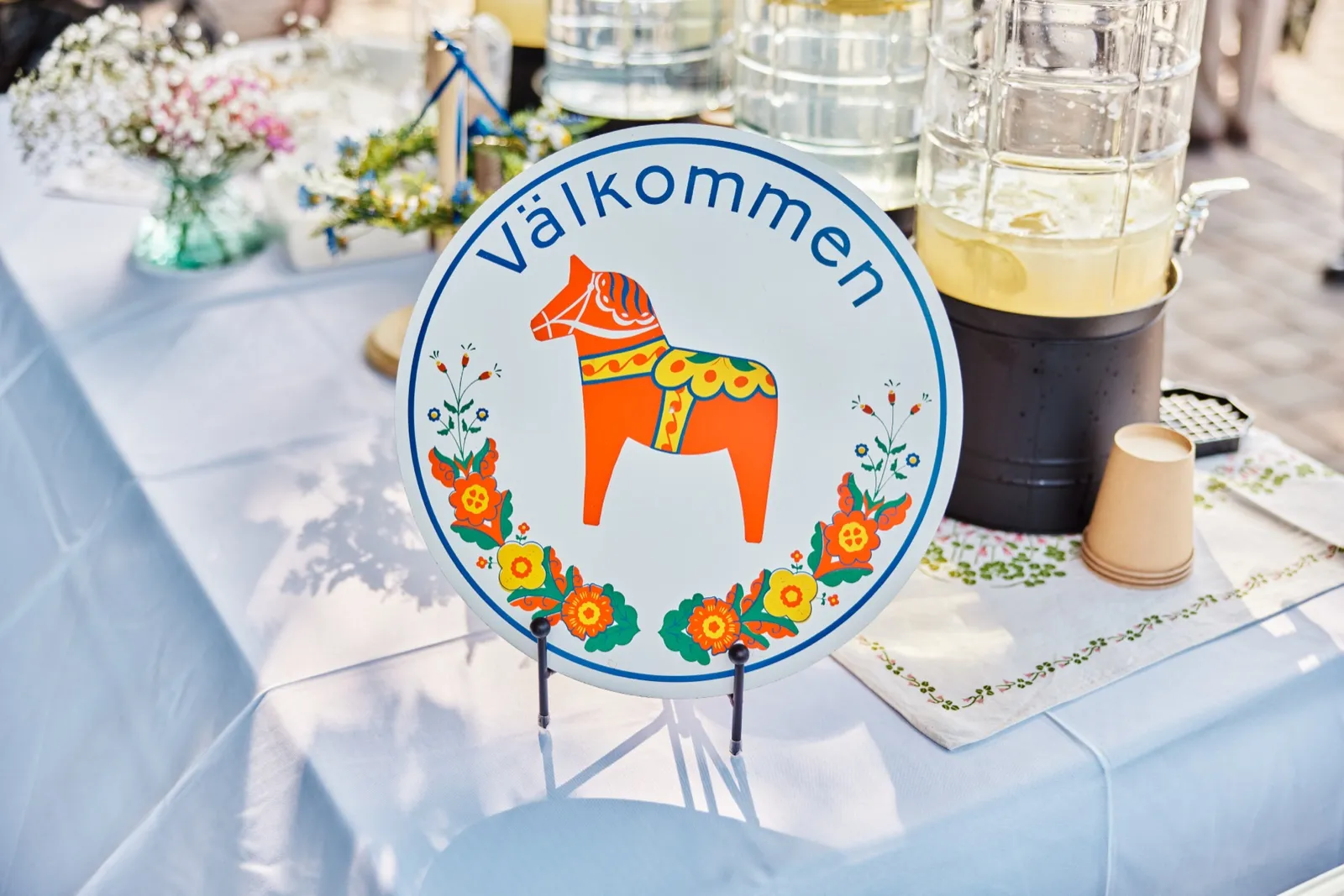 A round Swedish Välkommen sign painted with an orange Dala horse sits on a tablecloth-covered table beside lemonade dispensers and a small bouquet of wildflowers. — Tim Larsen Photography, Minnesota
