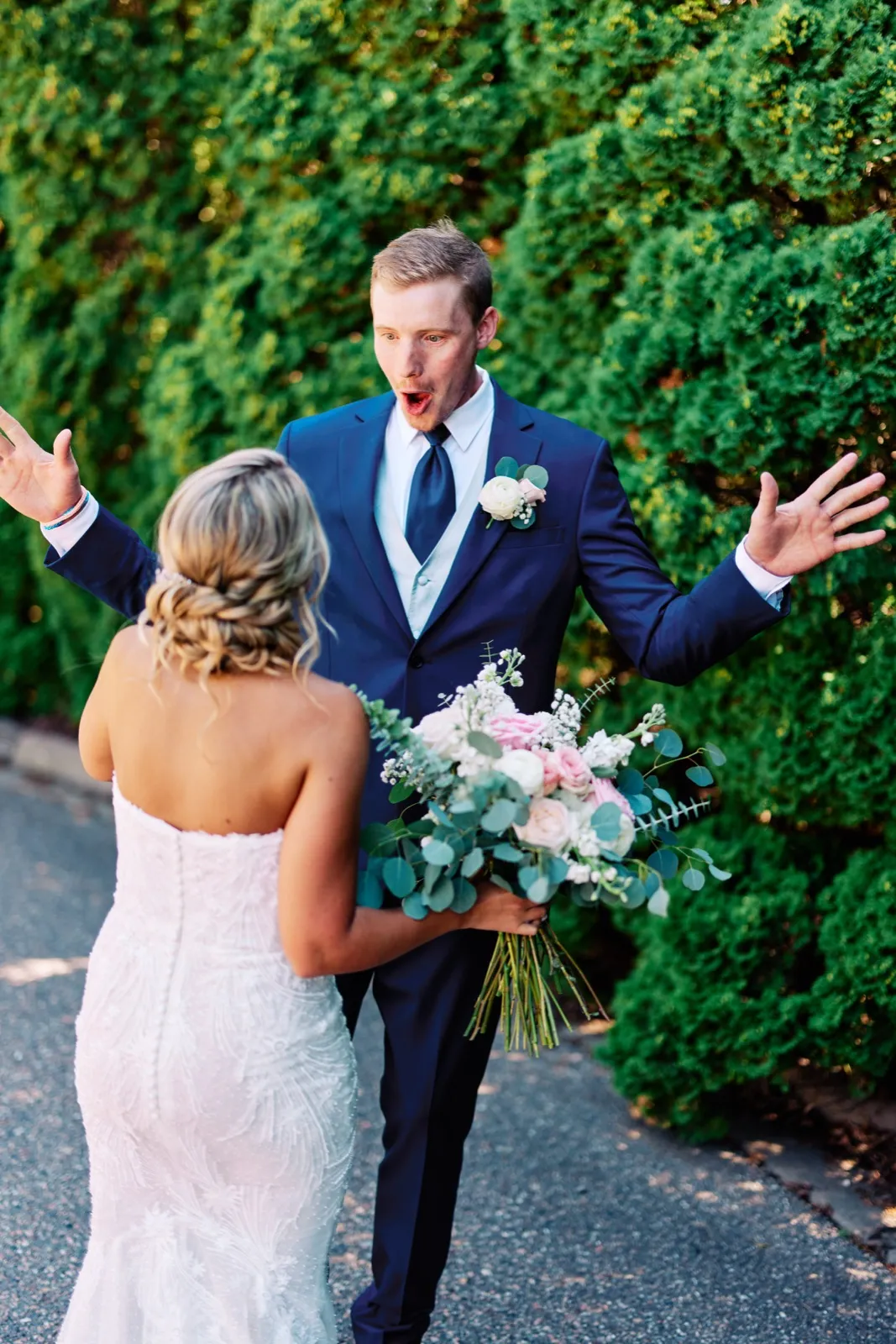 The groom throws his arms wide with a shocked, open-mouthed expression as he sees the bride for the first time on a garden path lined with tall evergreen hedges. — Tim Larsen Photography, Twin Cities MN
