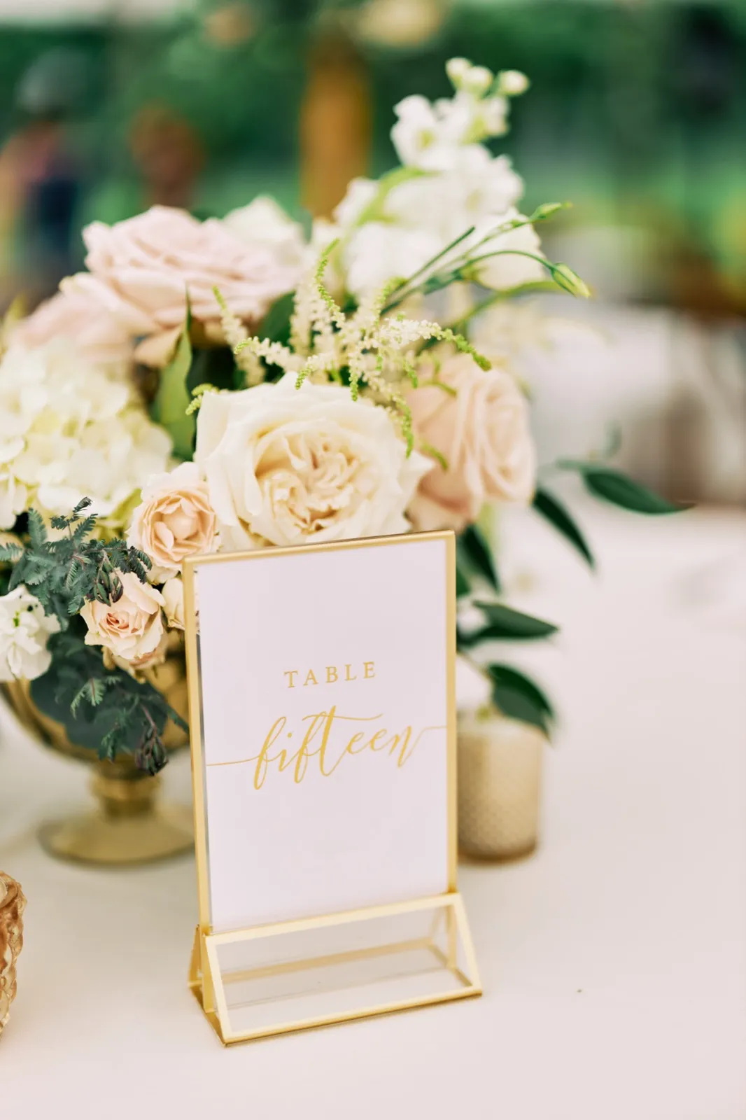 A gold-framed Table Fifteen sign sits beside a blush and ivory centerpiece of garden roses, hydrangea, and astilbe on a reception table. — Tim Larsen Photography, Brainerd Lakes MN