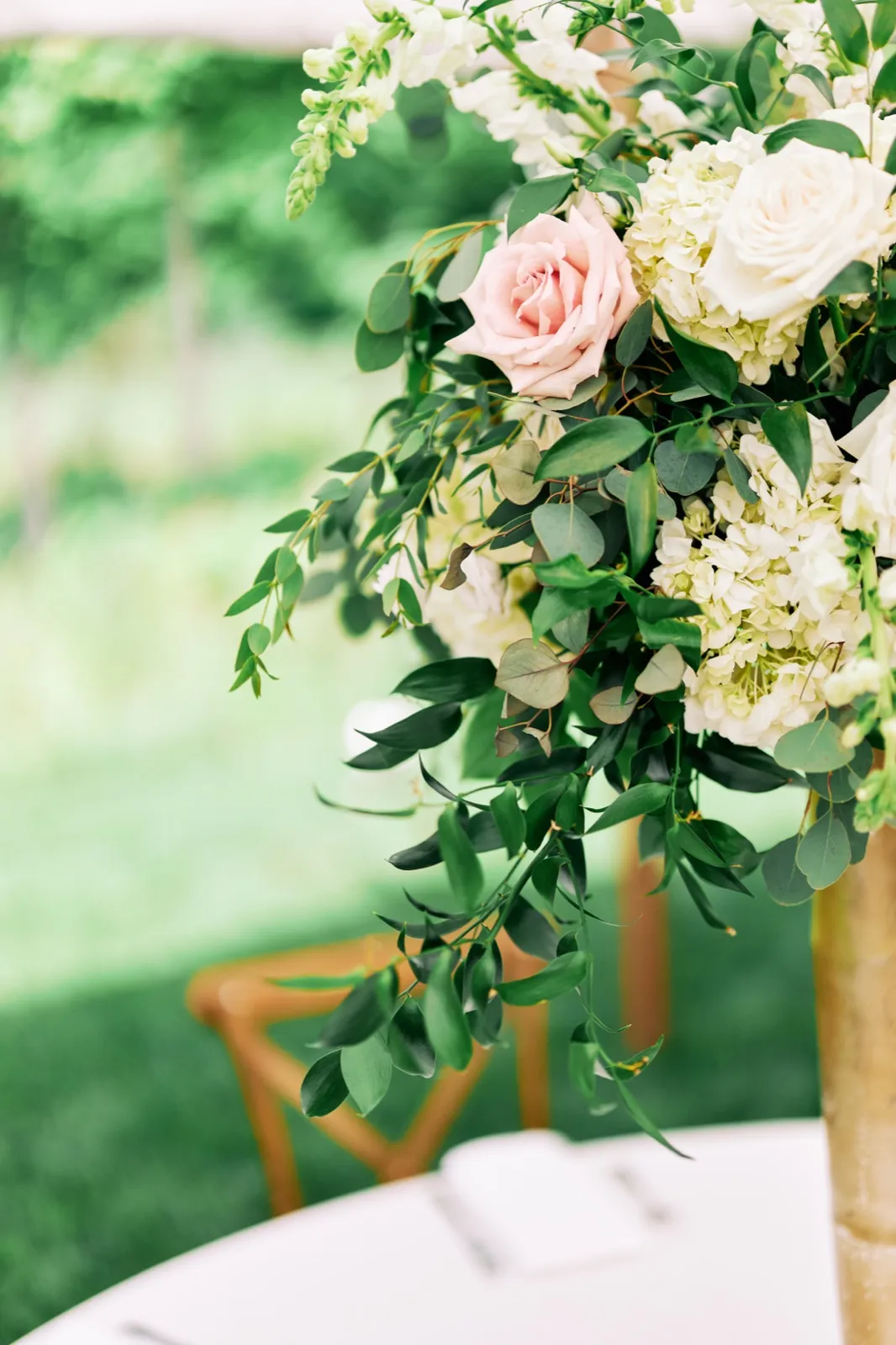 A tall reception centerpiece with a single blush rose, white hydrangeas, and trailing greenery sits on a table beside a wooden chair under a tent. — Tim Larsen Photography, Brainerd Lakes MN