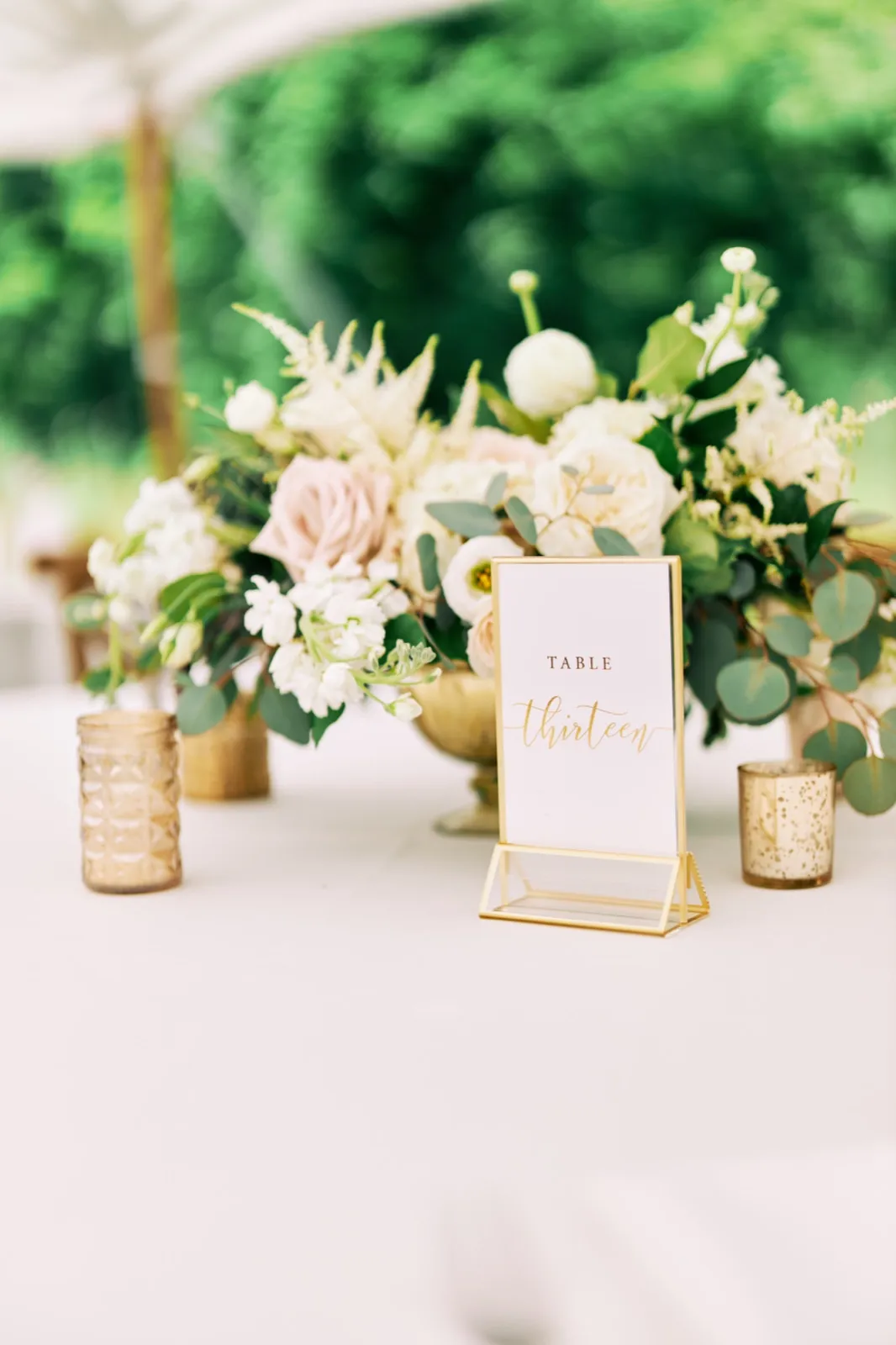 A gold-framed 'Table Thirteen' sign sits in front of a blush and white floral centerpiece with gold mercury votives on a tented reception table. — Tim Larsen Photography, Brainerd Lakes MN
