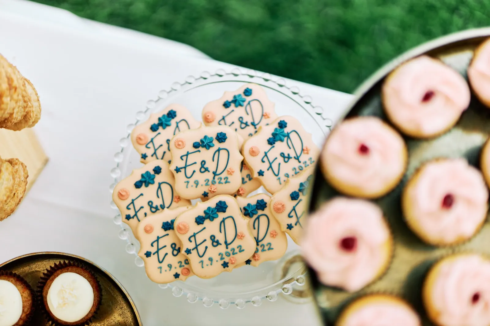 An overhead view of a glass cake stand holding iced sugar cookies monogrammed with 'E & D' and the date 7.9.2022, flanked by croissants and pink-frosted cupcakes. — Tim Larsen Photography, Brainerd Lakes MN