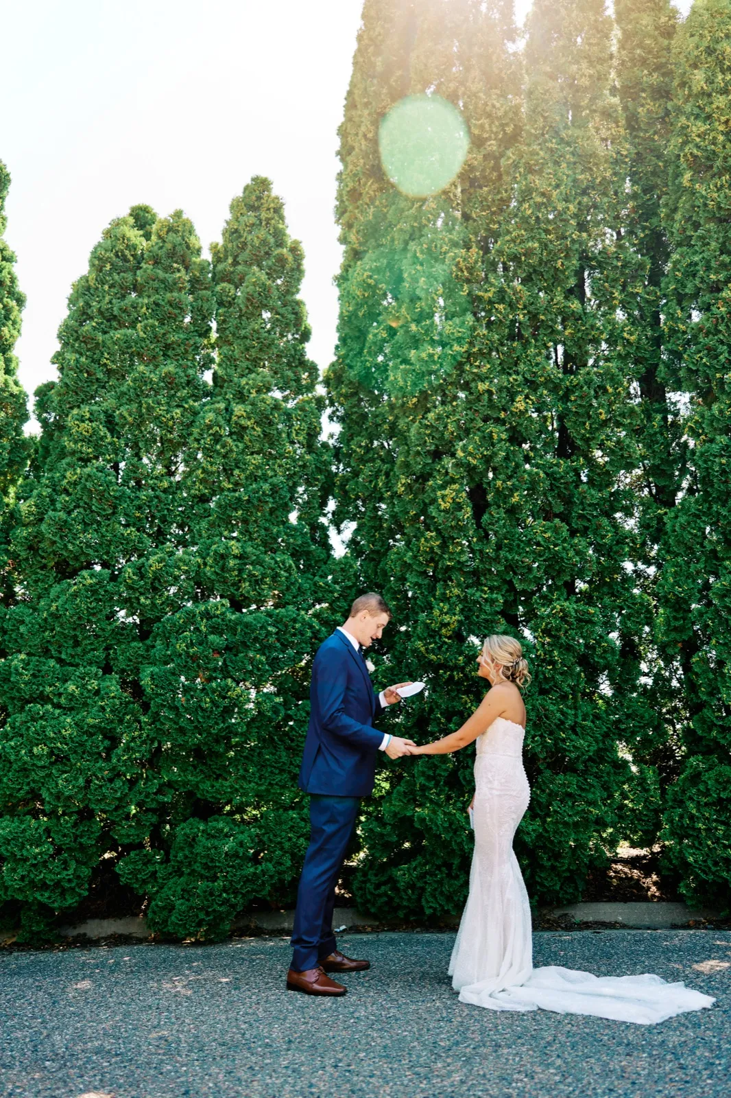 The groom reads his vows from a folded paper while holding the bride's hand, both standing on a paved path in front of a tall arborvitae hedge backlit by the sun. — Tim Larsen Photography, Twin Cities MN