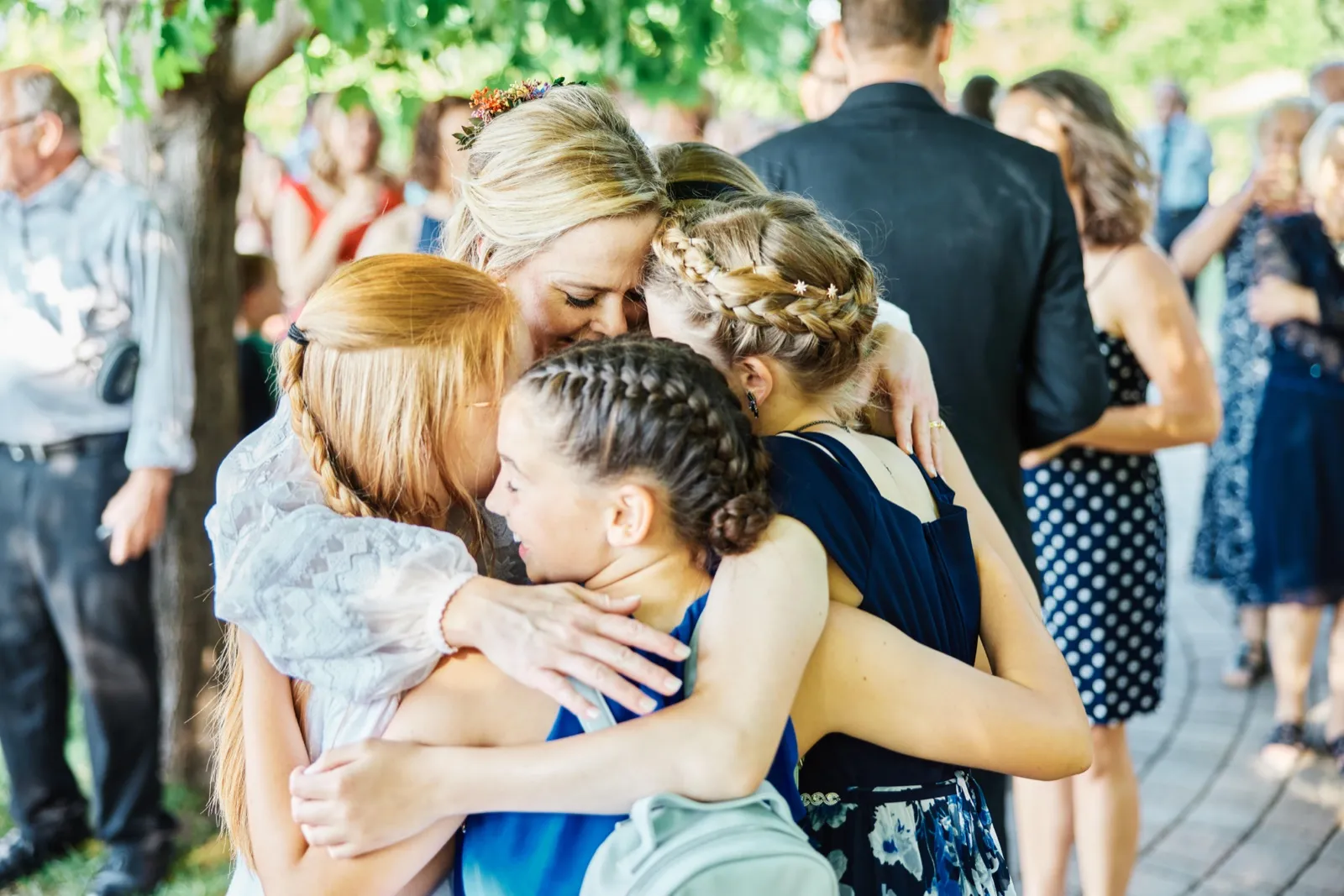The bride wearing a floral crown leans her forehead down into a tight group hug with three young girls in braids beneath dappled tree light after the ceremony. — Tim Larsen Photography, Minnesota