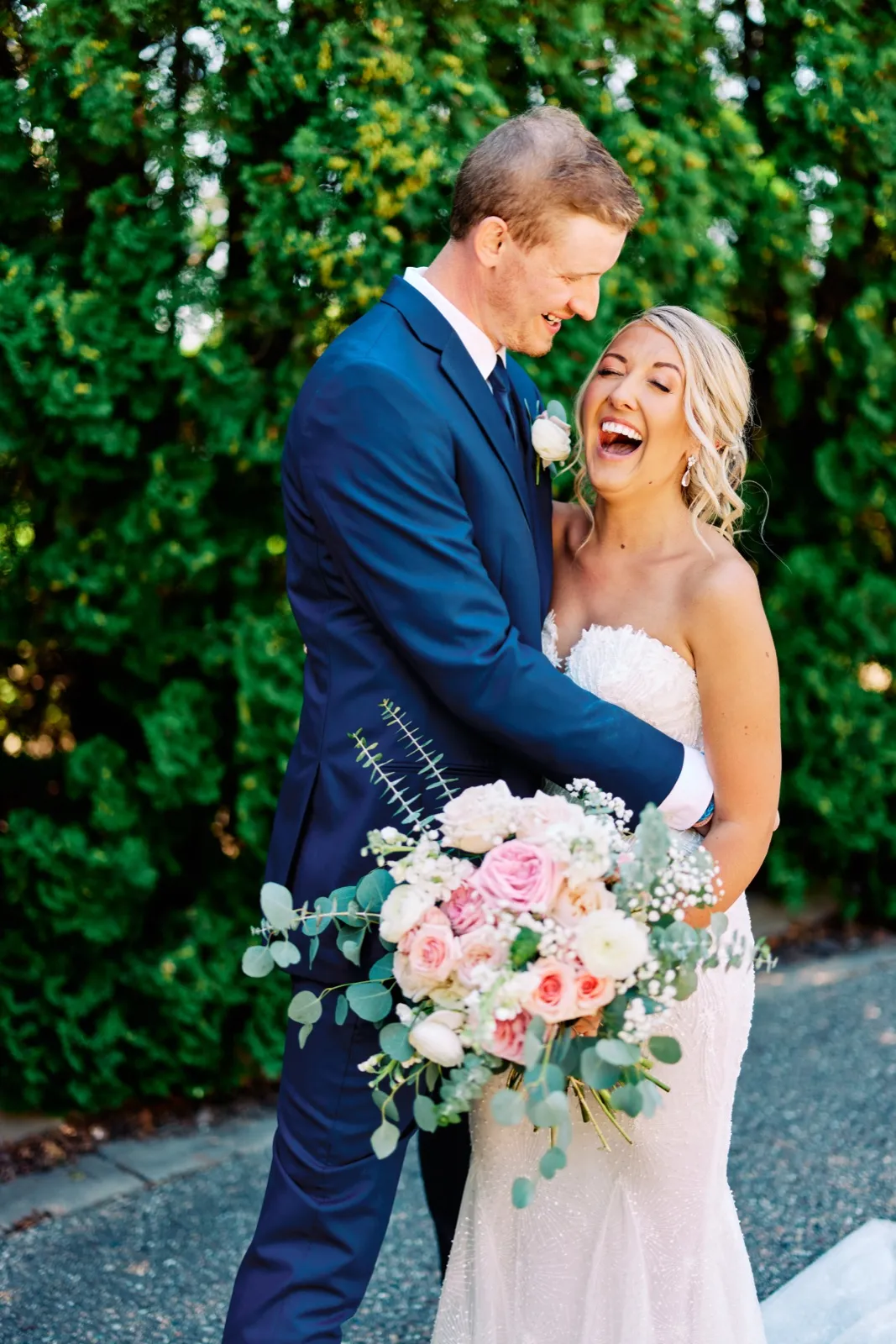 The bride throws her head back in open-mouthed laughter while the groom grins down at her, holding a pastel bouquet of roses and baby's breath against a tall green hedge. — Tim Larsen Photography, Twin Cities MN
