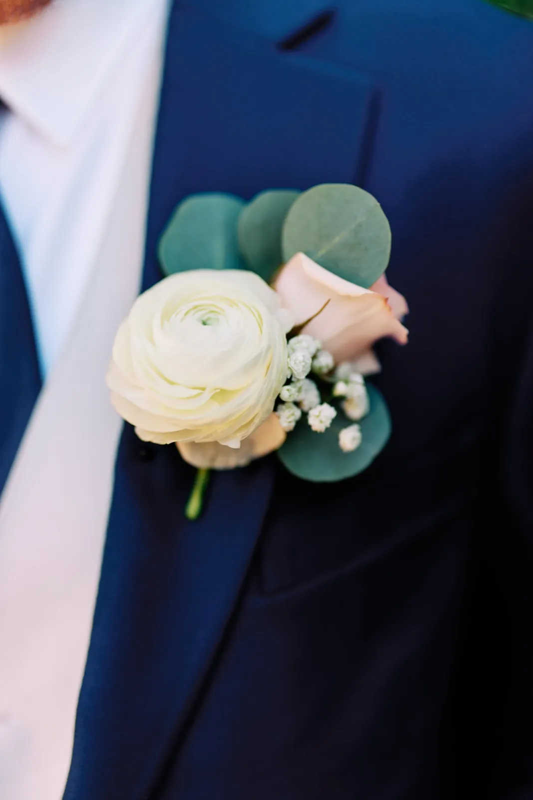 A close-up of the groom's boutonniere — a white ranunculus paired with a blush rose, baby's breath, and eucalyptus pinned to a navy suit lapel. — Tim Larsen Photography, Twin Cities MN