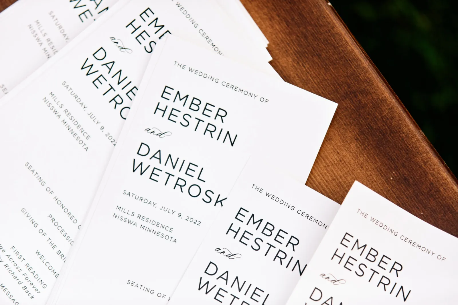 A stack of wedding ceremony programs for Ember Hestrin and Daniel Wetroski, dated July 9, 2022, fanned across a dark wood surface. — Tim Larsen Photography, Brainerd Lakes MN