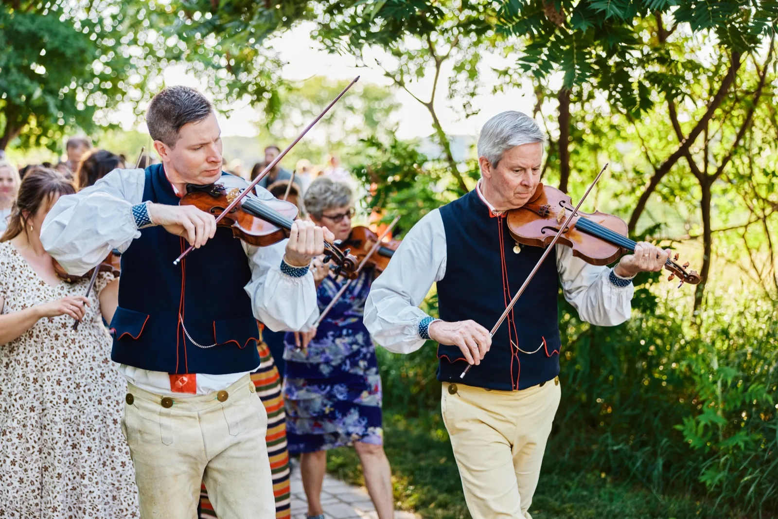 Two violinists in traditional embroidered vests play as they lead a procession of guests down a sunlit garden path. — Tim Larsen Photography, Minnesota