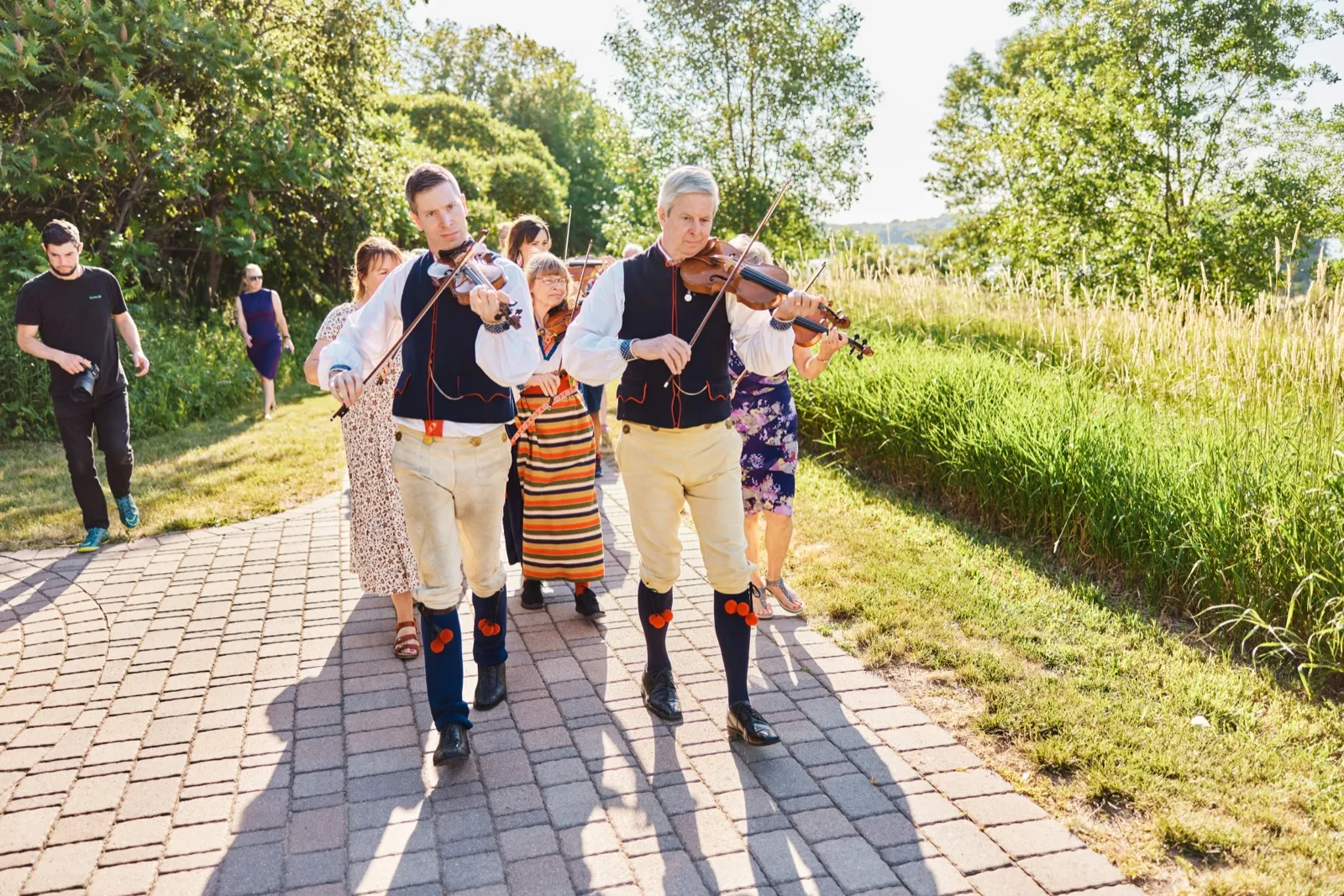 Musicians in traditional Scandinavian folk costume play violins as they walk down a brick path in evening sunlight, followed by guests and another photographer. — Tim Larsen Photography, Minnesota
