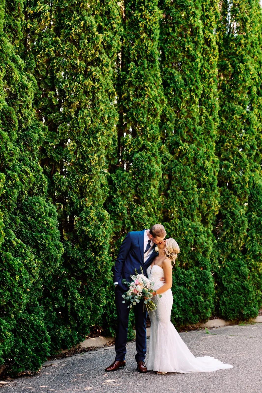 The bride and groom share a kiss on a paved path beside a tall arborvitae hedge that towers above them, the bride holding a pastel bouquet. — Tim Larsen Photography, Twin Cities MN