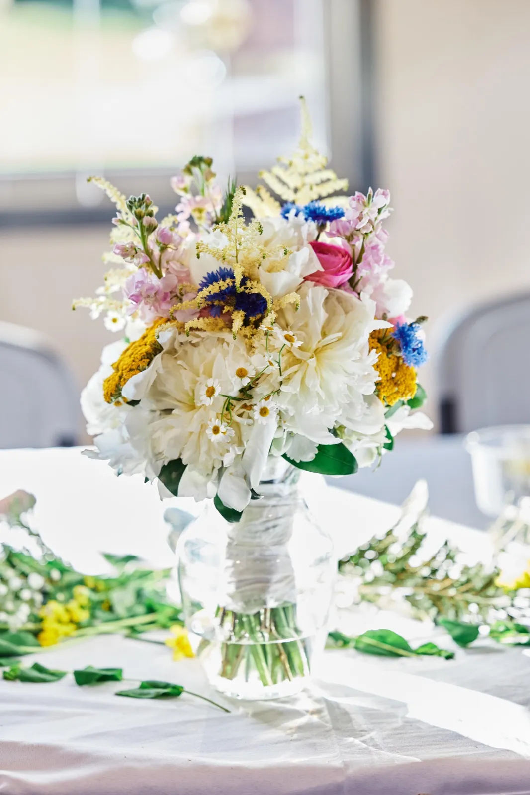 A wildflower bridal bouquet with white peonies, blue cornflowers, pink stock, and yellow yarrow sits in a glass vase on a reception table near a window. — Tim Larsen Photography, Minnesota