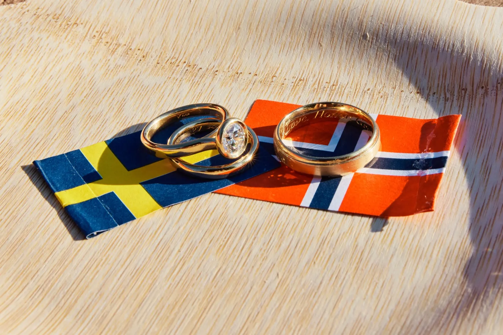 Three gold wedding rings, one with a diamond, rest on small Swedish and Norwegian flags atop a sunlit wooden surface. — Tim Larsen Photography, Minnesota