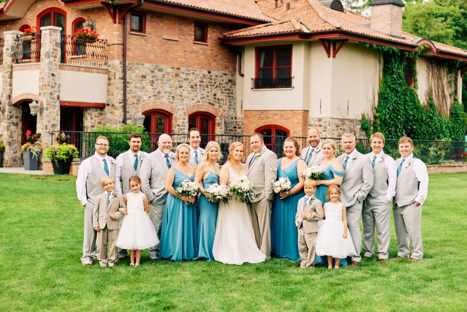 The bride and groom stand center with their full bridal party, flower girls, and ring bearers on a lawn in front of a large stone-and-brick lodge. — Tim Larsen Photography, Brainerd Lakes MN