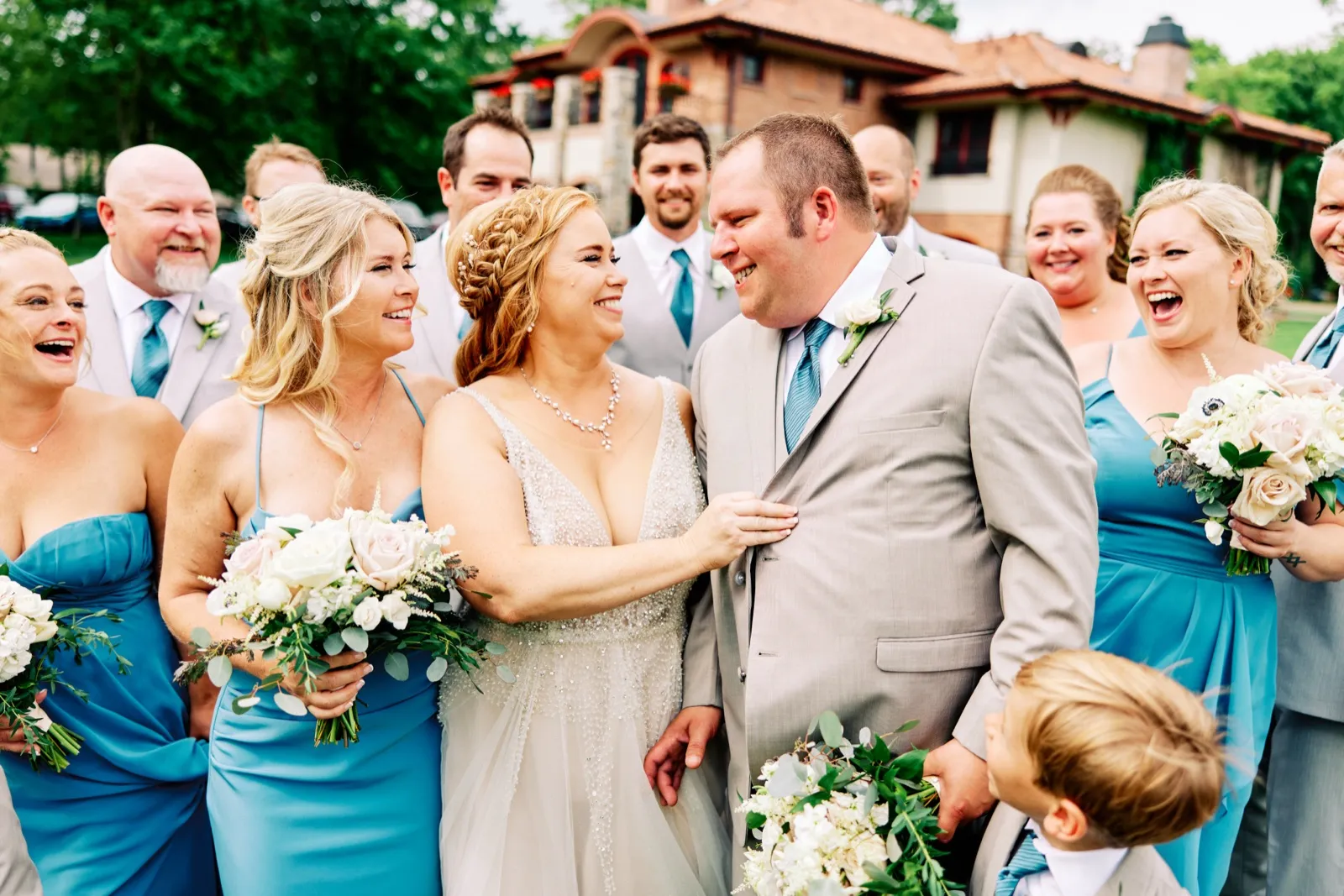 The bride and groom laugh face-to-face surrounded by their bridal party in dusty-blue dresses and gray suits on a lawn outside a brick lodge. — Tim Larsen Photography, Brainerd Lakes MN