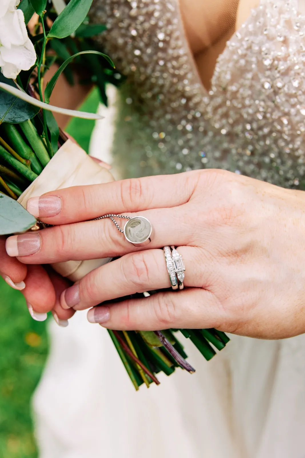 The bride's hands cradle her bouquet, displaying her stacked diamond rings alongside a small round memorial locket pendant resting against her beaded gown. — Tim Larsen Photography, Brainerd Lakes MN
