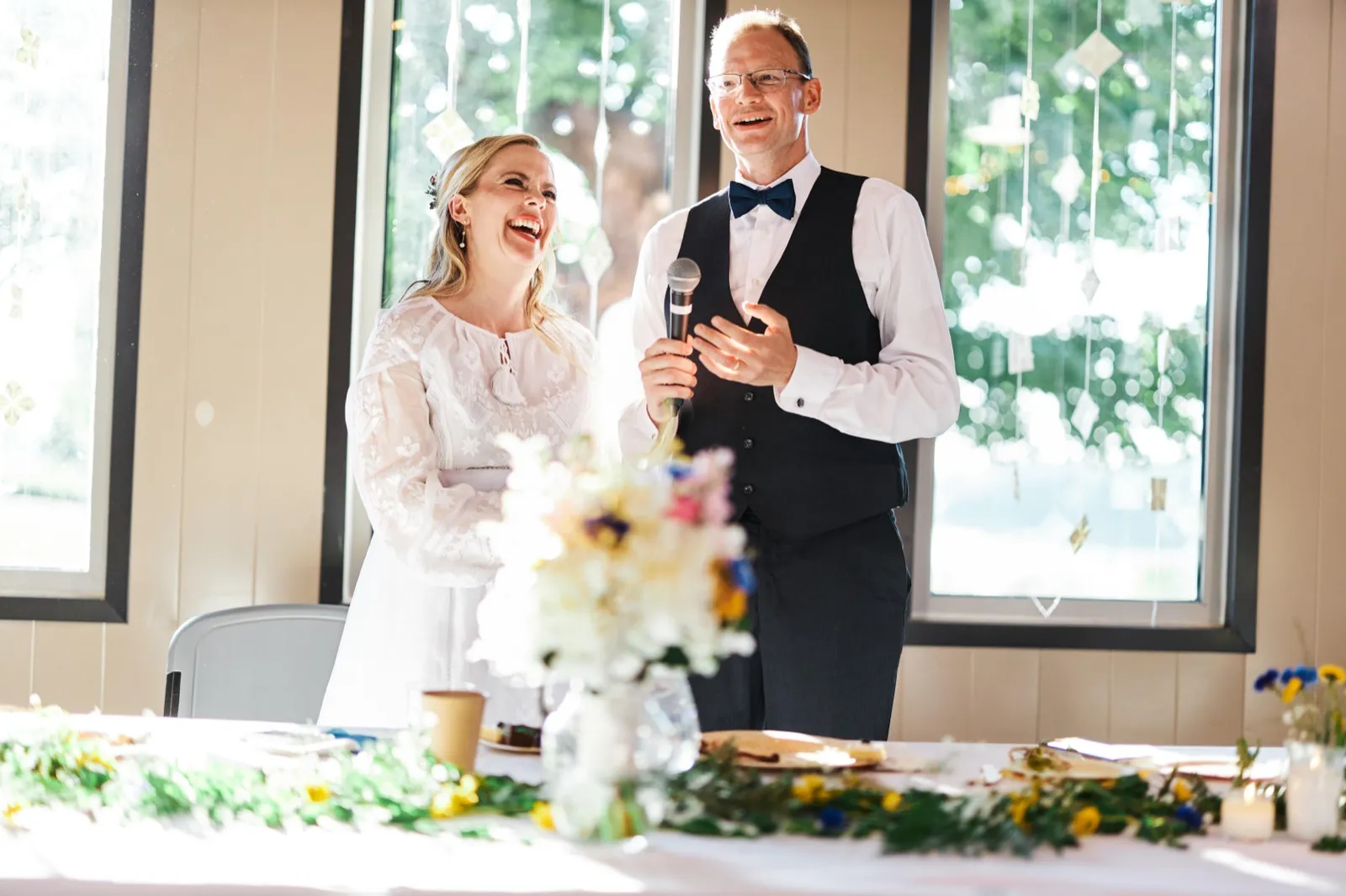 The bride laughs openly beside the groom as he holds a microphone and addresses guests during a reception toast in front of bright windows. — Tim Larsen Photography, Minnesota